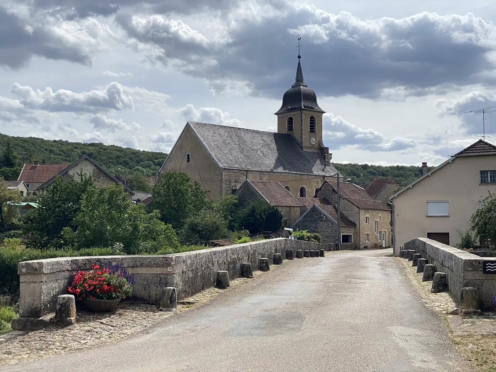 A stone humpback bridge decked with red flowers leads into a village beneath a tall church bell tower with a slate spire, hills wooded behind.