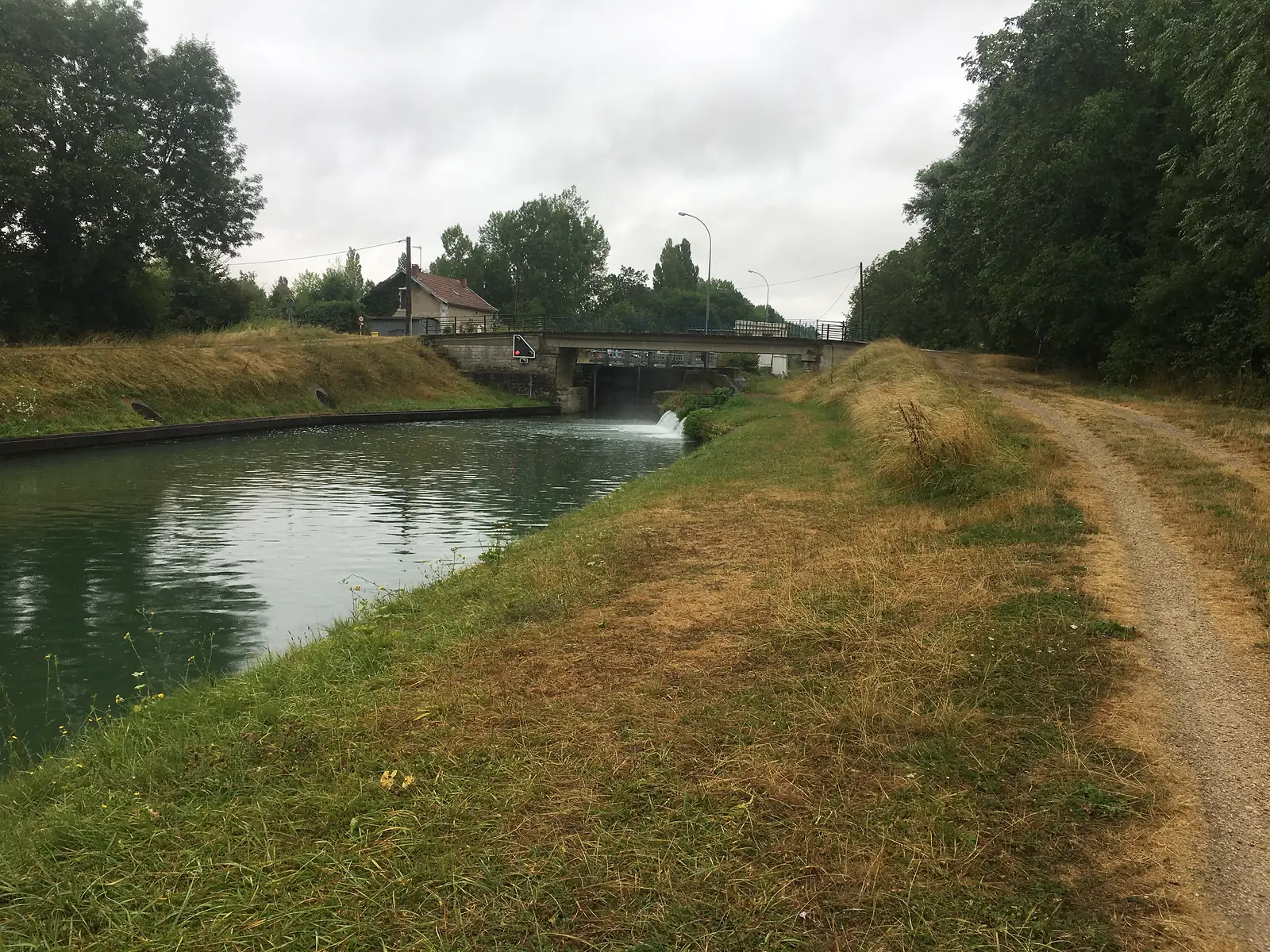A canal lock with water spilling beneath a road bridge, a house and street lamps visible beyond, with a grassy towpath curving away to the right.