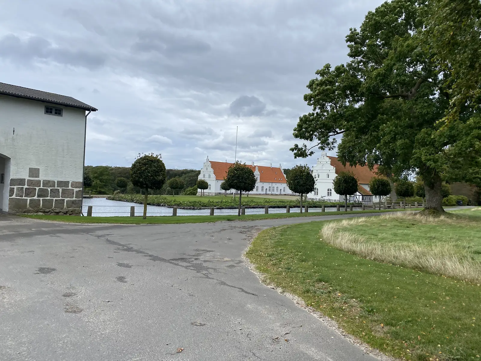 A white crow-stepped manor with a red-tiled roof beyond its moat and a row of clipped standard trees, seen from a lane past a rendered outbuilding.