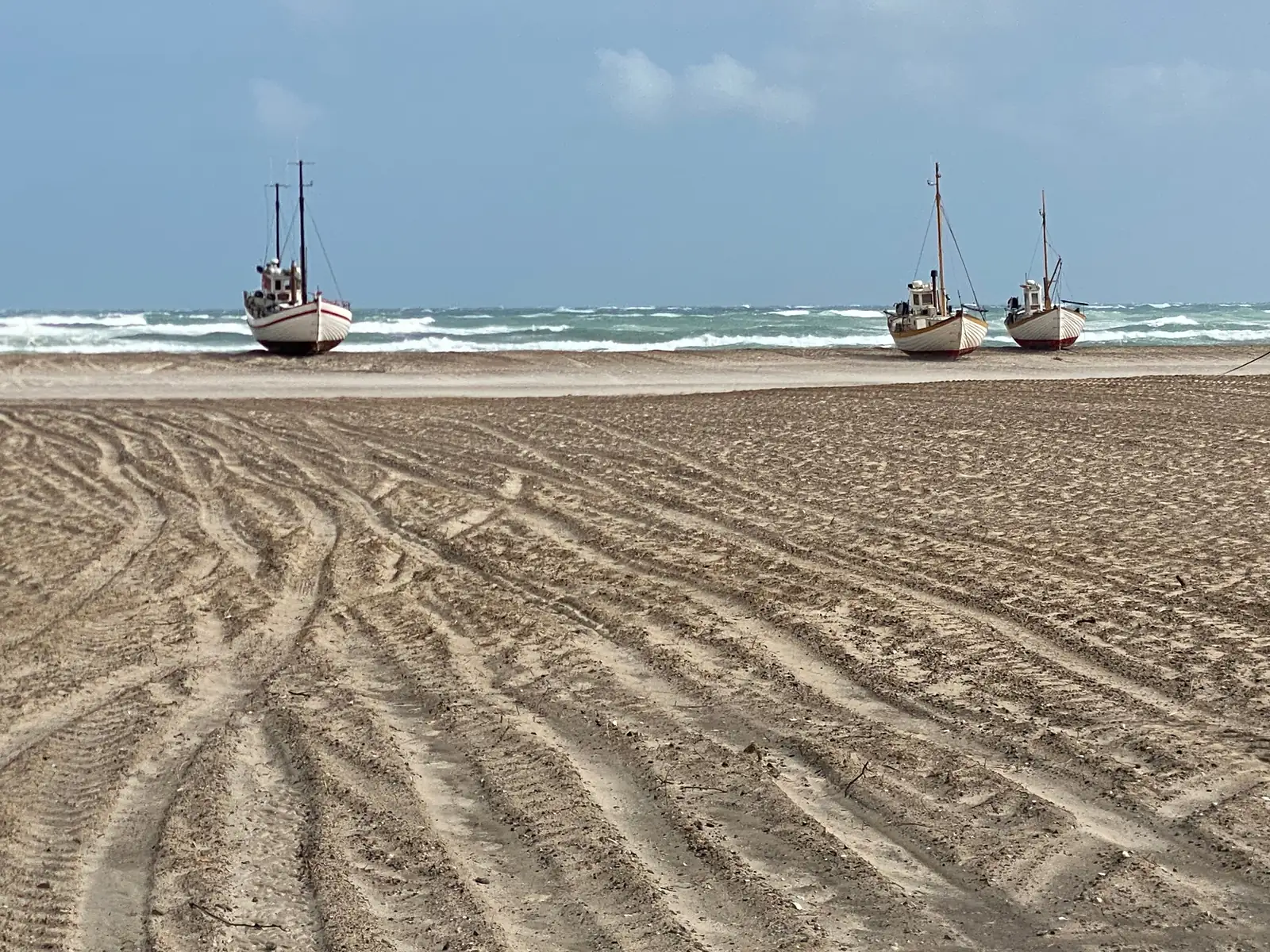 Three white beach-launched fishing boats at the water's edge beyond a wide expanse of sand deeply combed with tractor tracks.