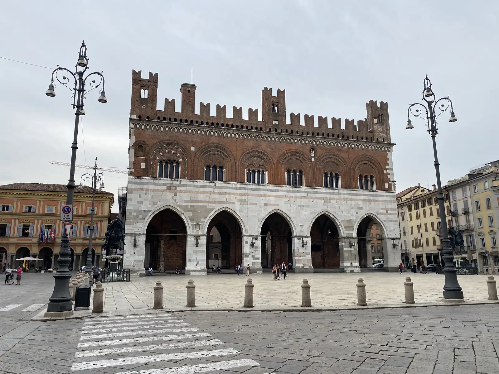 The crenellated brick and marble facade of the Palazzo Gotico rises above an empty piazza in Piacenza, its Gothic arcade open onto the square below.