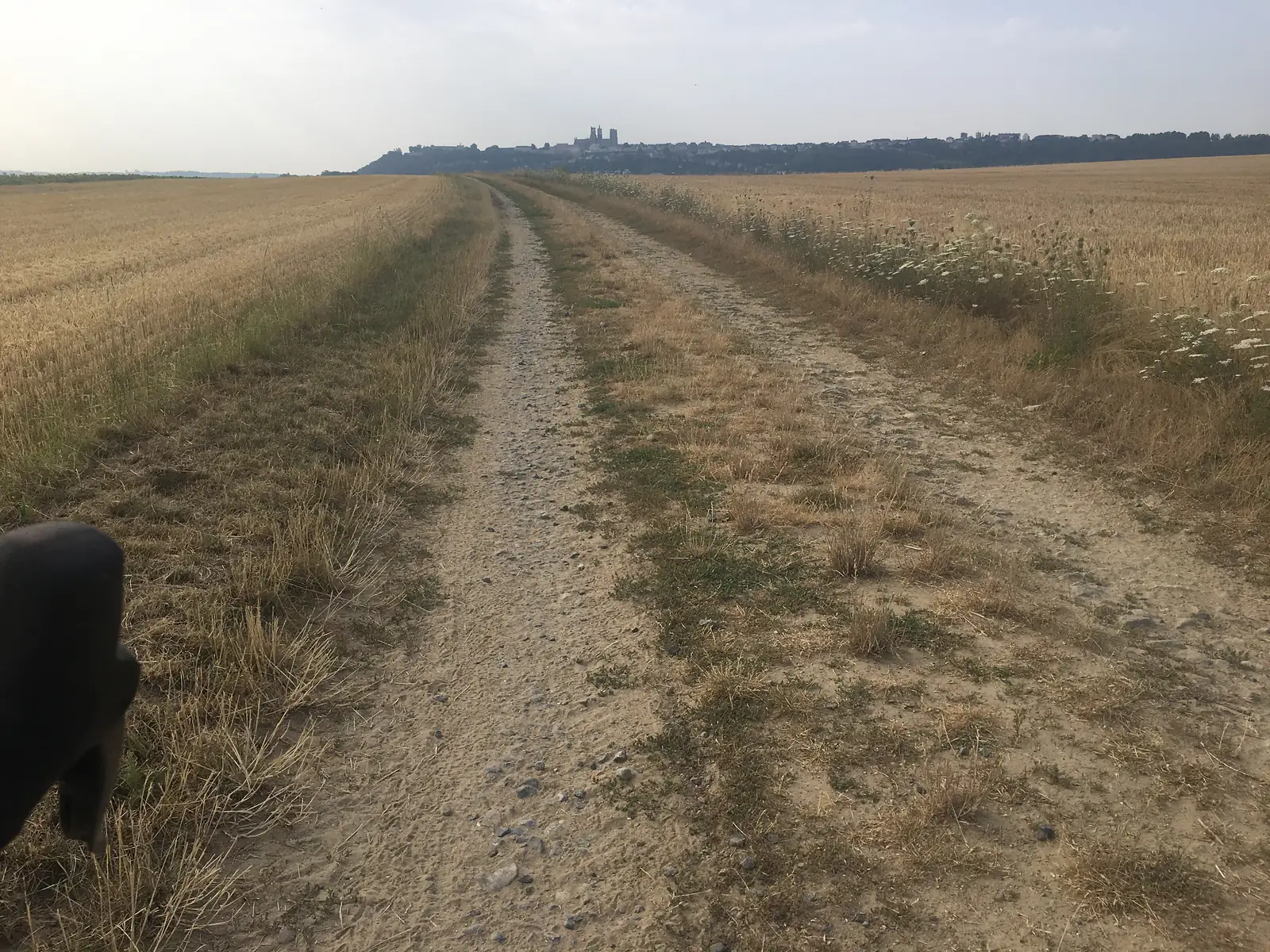 A dusty farm track leads towards a hilltop town whose cathedral towers stand out clearly on the skyline, a dark pannier bag visible in the foreground.