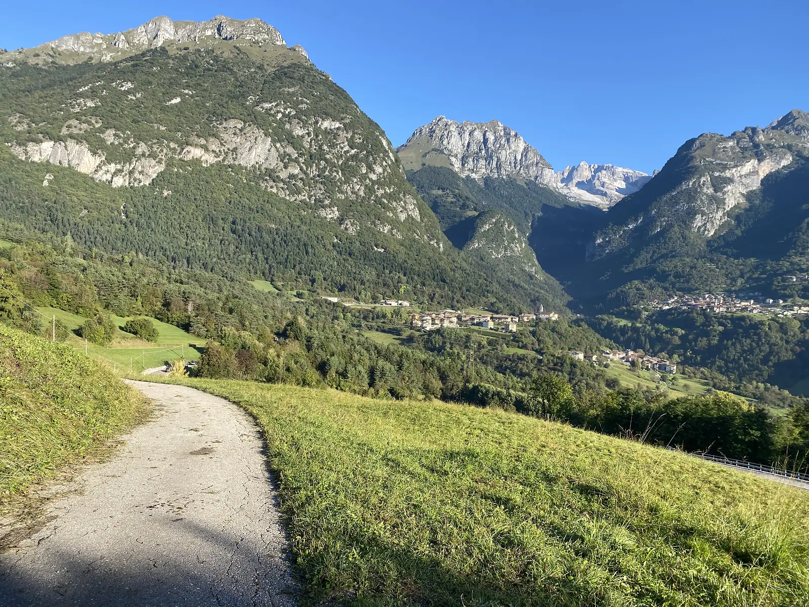 A tarmac lane descends through meadows towards a village strung along the valley floor, with pale rocky peaks and a high snow-streaked ridge behind.