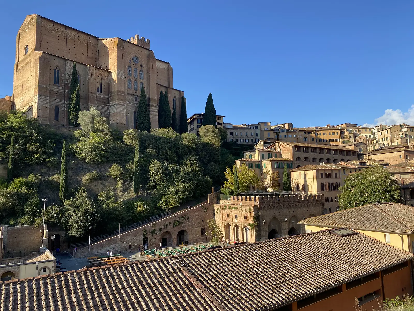 A large brick basilica crowning a Tuscan hilltop town, surrounded by cypress trees and terracotta rooftops under a clear sky.