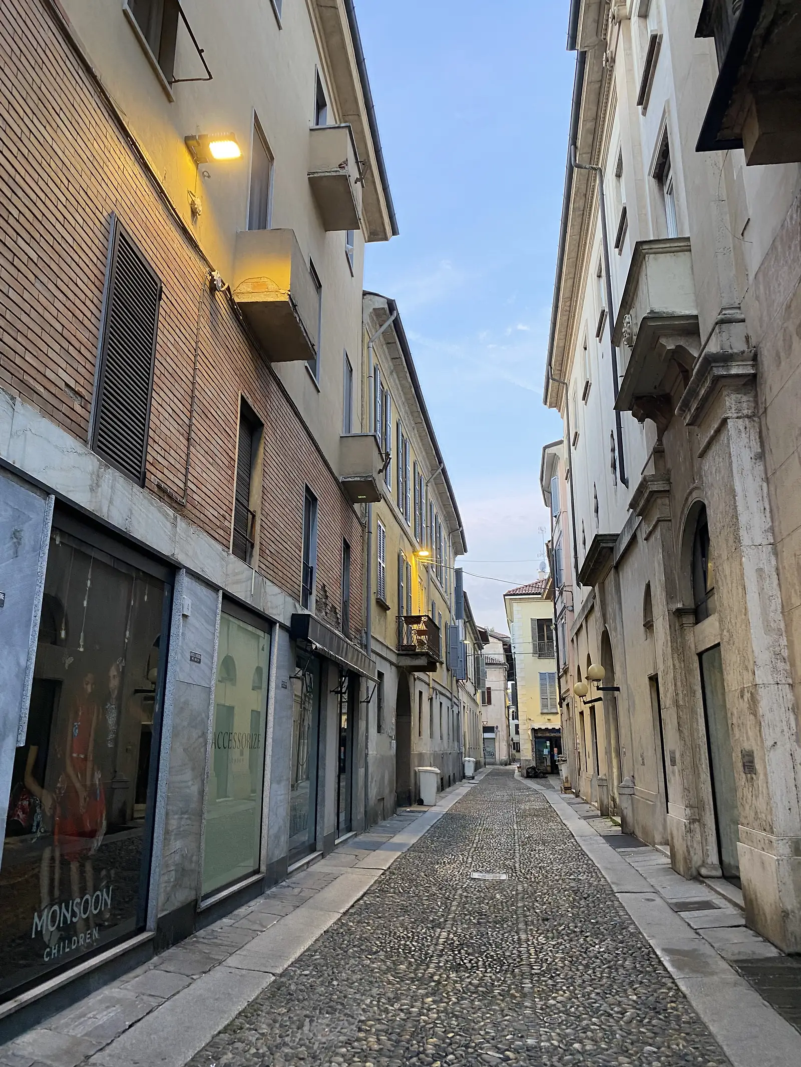 A narrow, pebble-paved street between arcaded stone buildings, with shopfronts for Accessorize and Monsoon Children beneath shuttered upper windows.