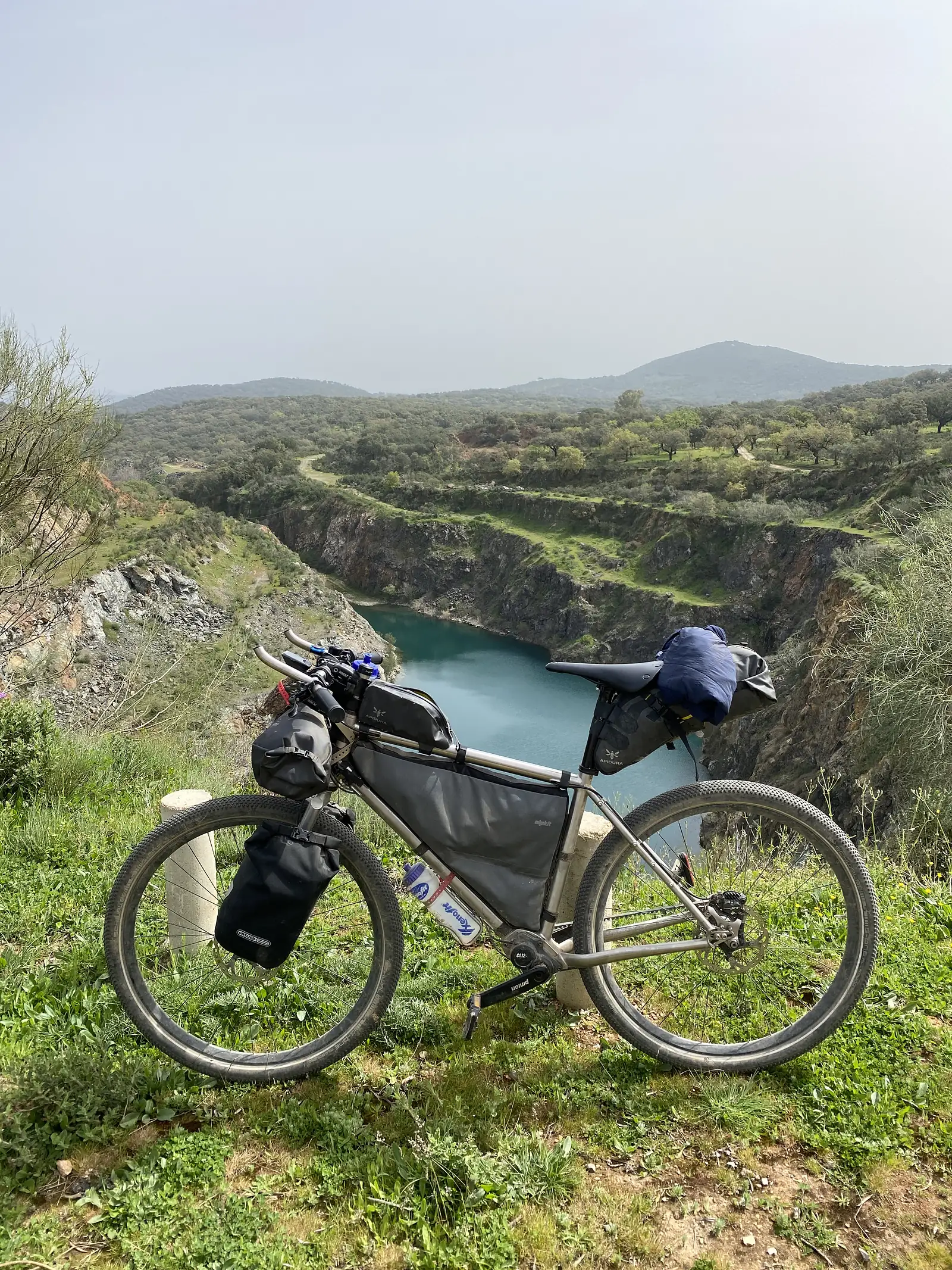 A loaded bikepacking bike propped on the grassy rim of a flooded quarry, its turquoise water ringed by rock ledges and oak pasture.