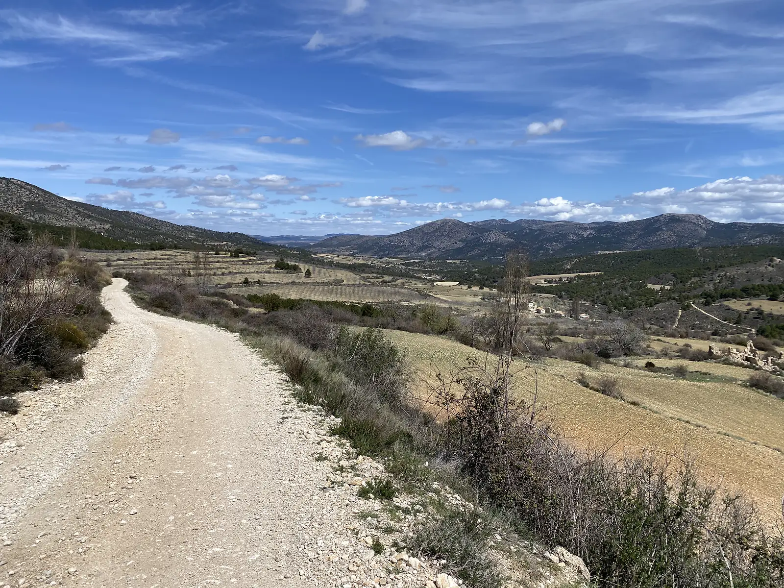 A gravel track curving down a hillside above a broad valley of ploughed fields, olive groves and a lone farmstead, ringed by mountains.