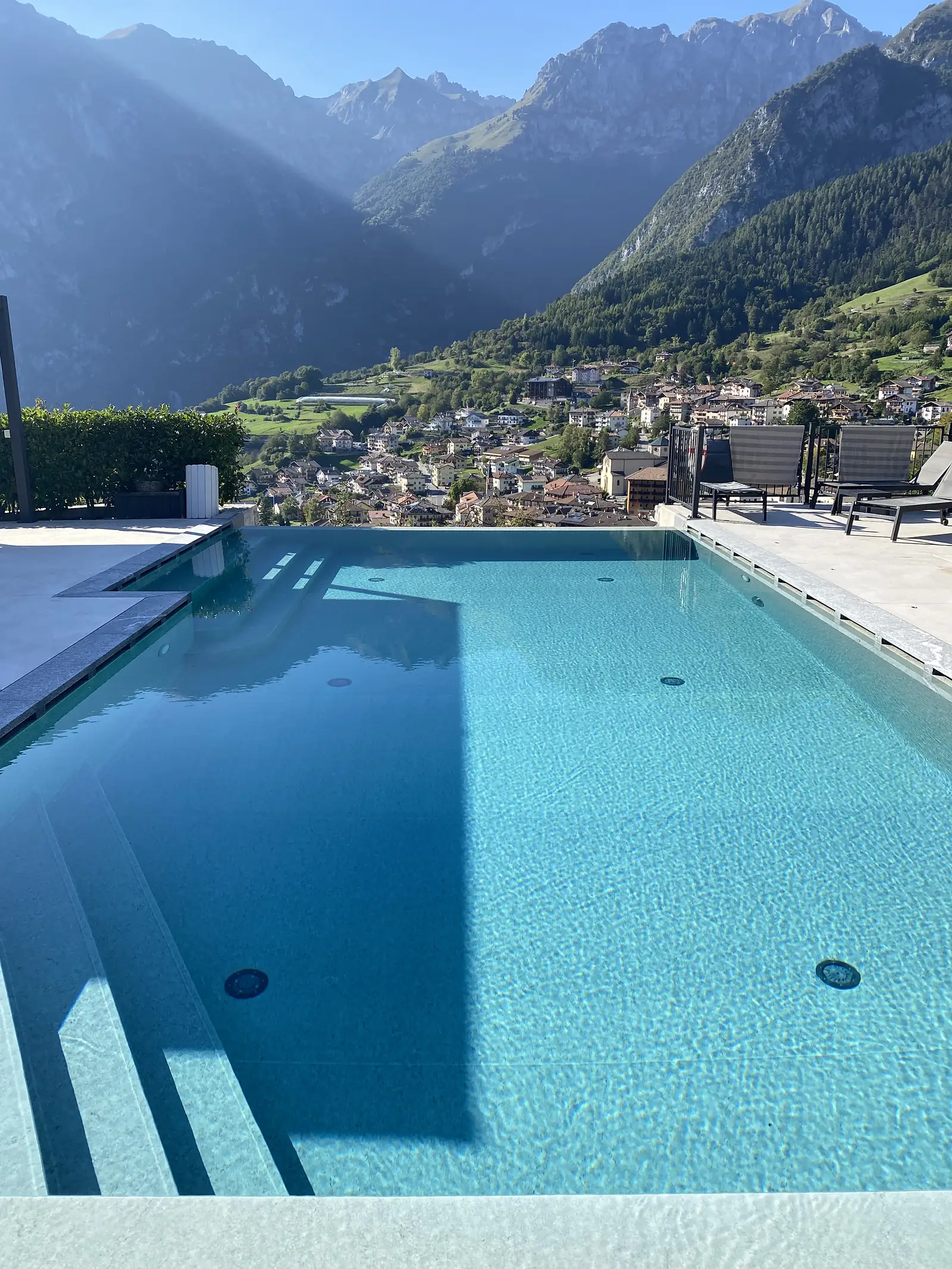 An infinity pool and sun loungers on a terrace look out over a village filling a steep green valley ringed by mountains.