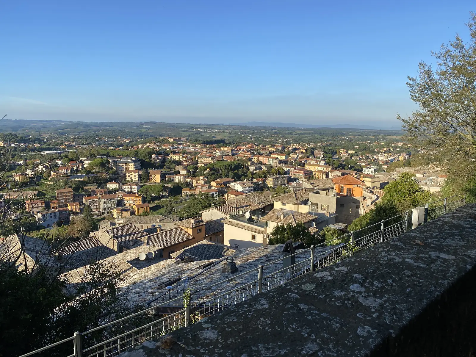Rooftops of a hilltop town spill down towards a wider modern town in the plain below, seen from a stone terrace.