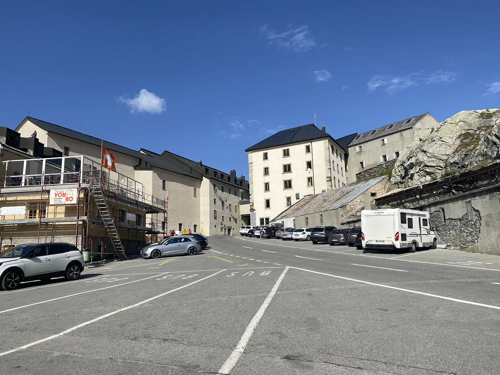 Cars and a camper van are parked outside a row of stone buildings with dark roofs at the mountain pass, one under scaffolding and marked 'Von Ro'.
