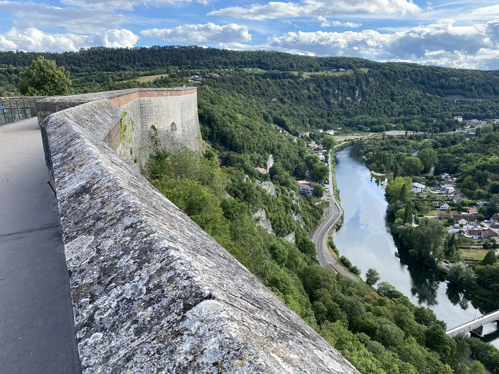 A rampart wall curves along a cliff edge above a wooded valley, with a stone bastion projecting out among the trees.