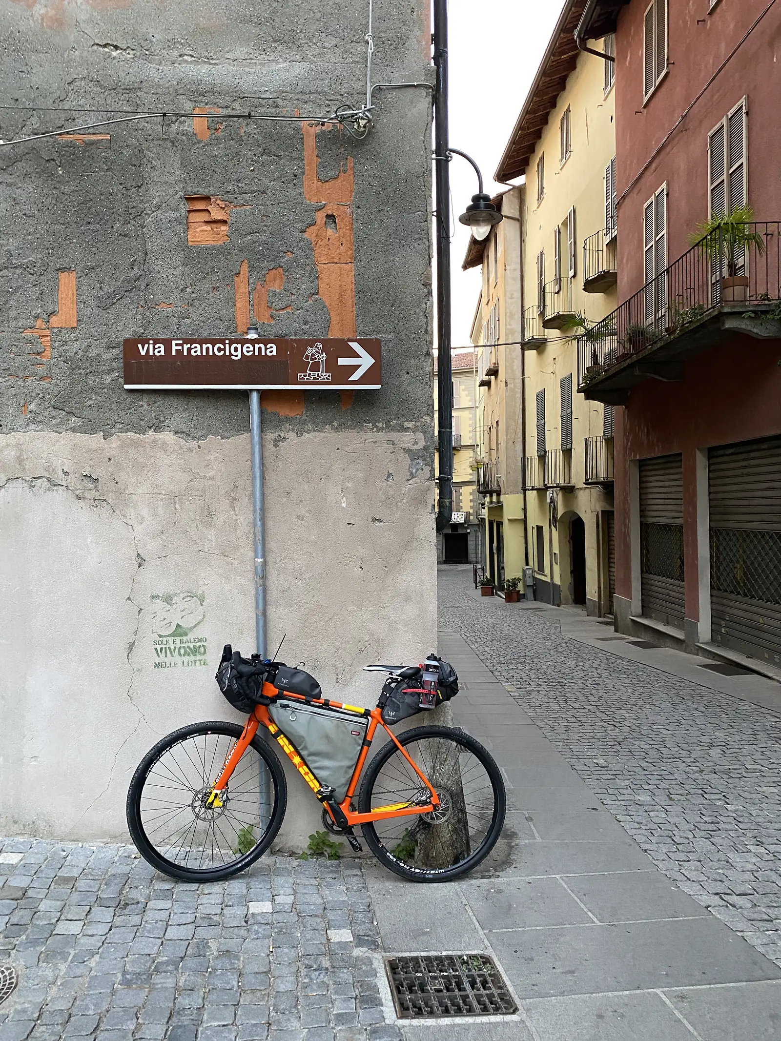 A laden orange gravel bike leaning against a cobbled wall beneath a brown Via Francigena waymarker pointing straight ahead, with pastel Italian townhouses lining the alley behind.