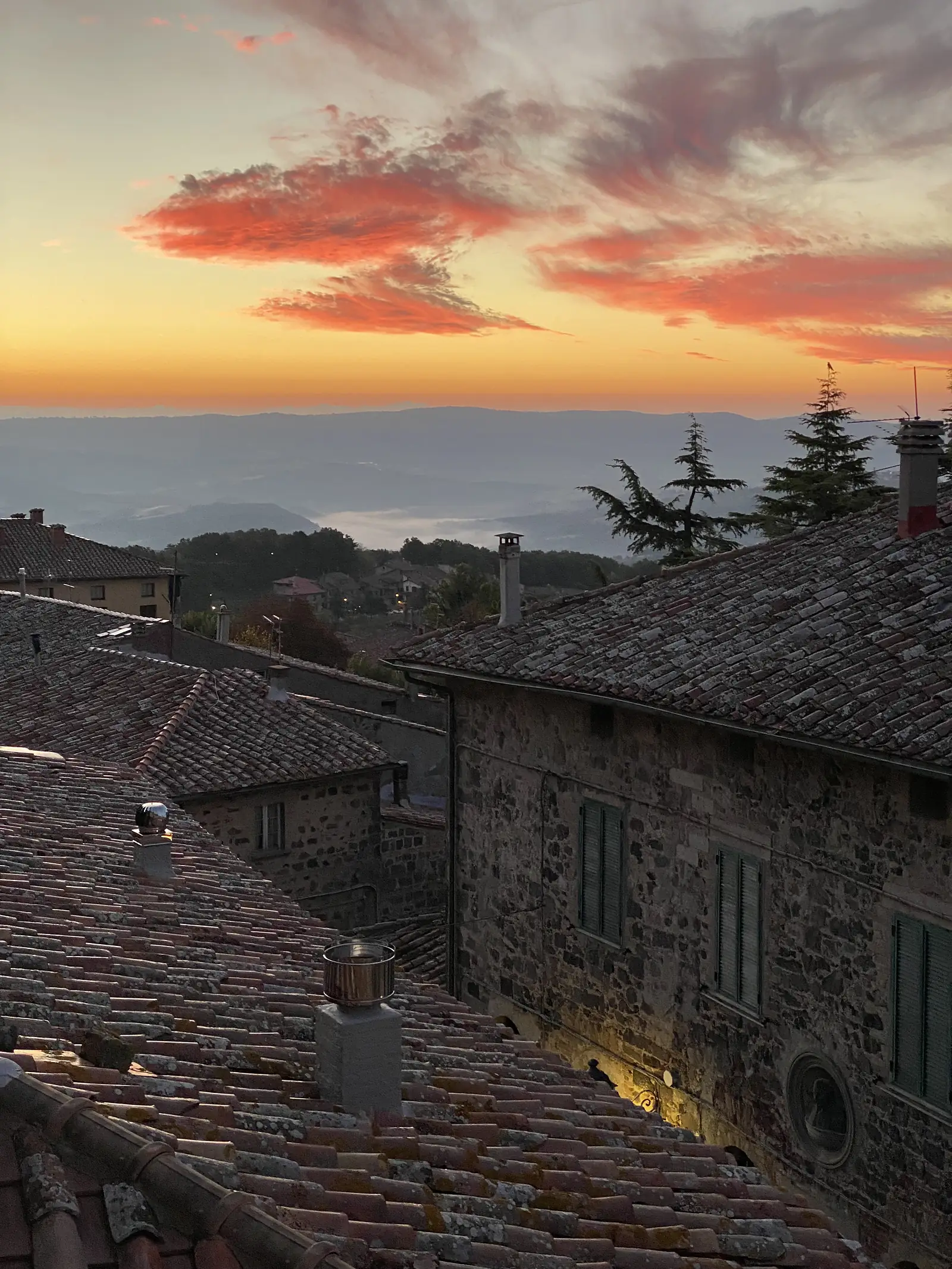 A fiery orange and red sunset sky over terracotta rooftops and a stone bell tower in a hilltop Italian town.