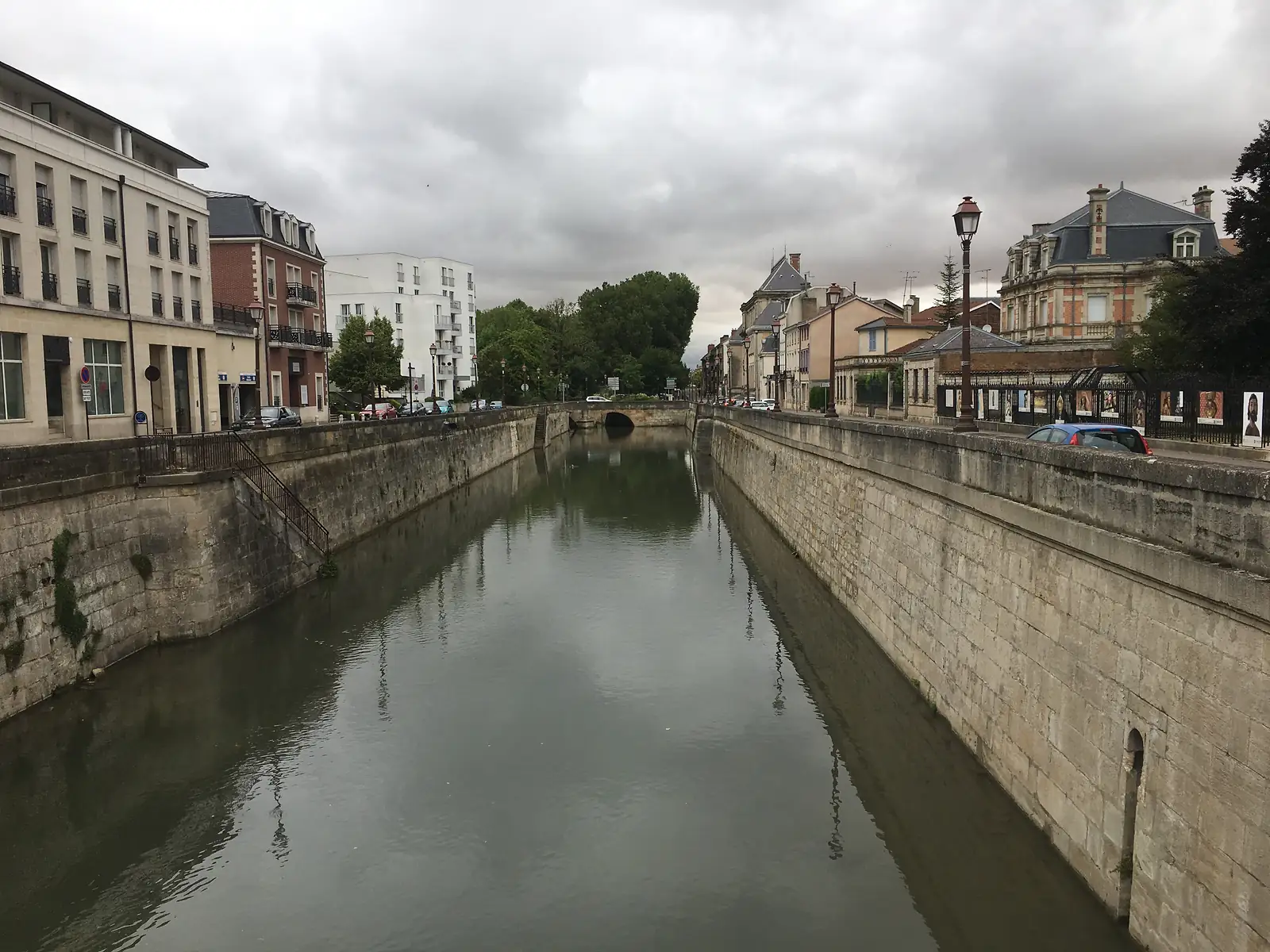 A stone-walled canal channel runs through the centre of Châlons-en-Champagne, flanked by parked cars, street lamps and a mix of period buildings under a cloudy sky.