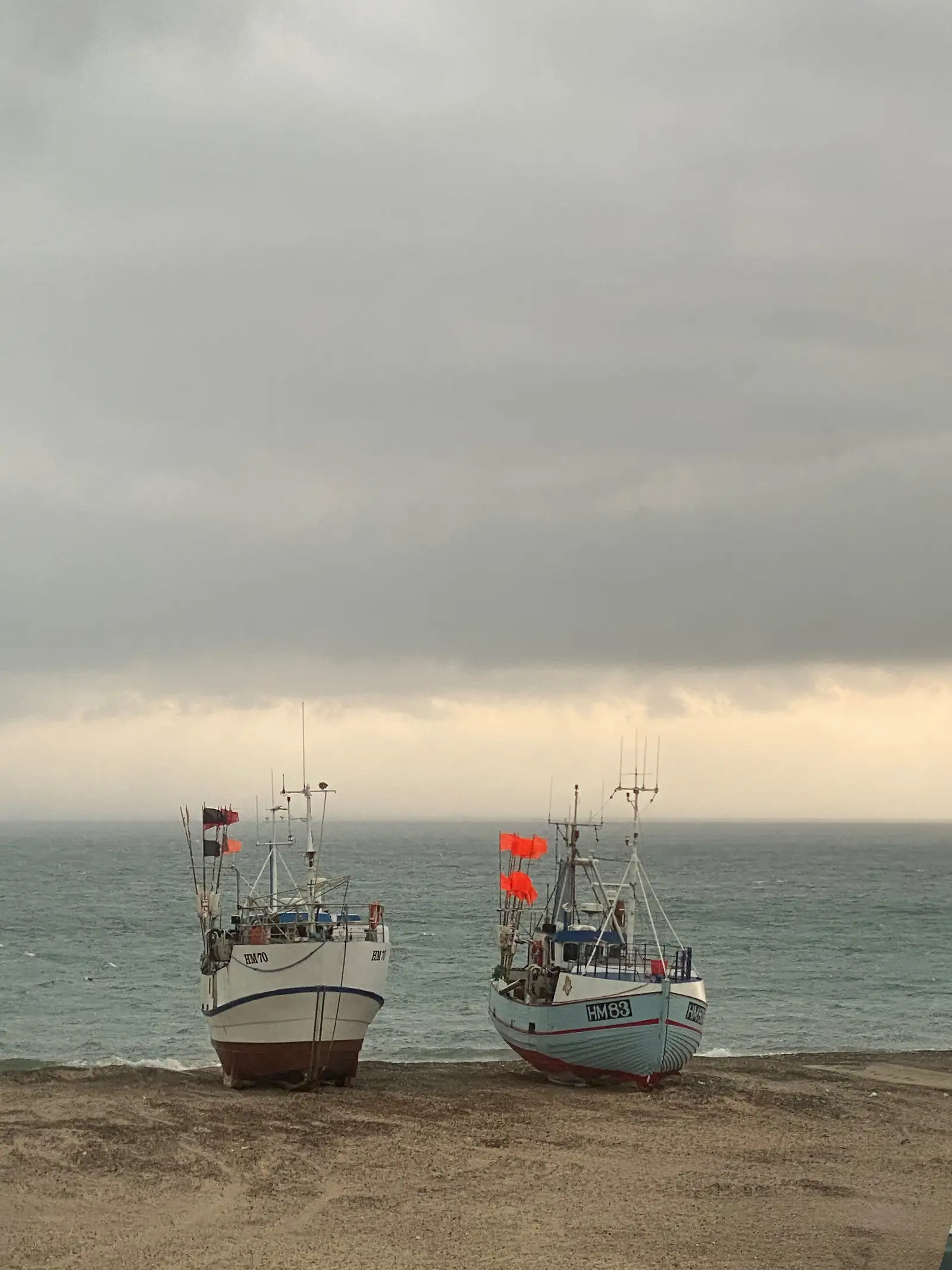 Two beach-launched fishing boats drawn up on the sand with orange marker flags, under a grey sky over a choppy sea.