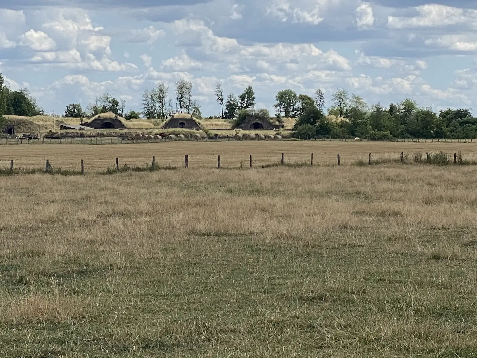 Traditional turf-roofed stone barns sit low in a stubble field with round hay bales scattered in front, fringed by a line of trees.