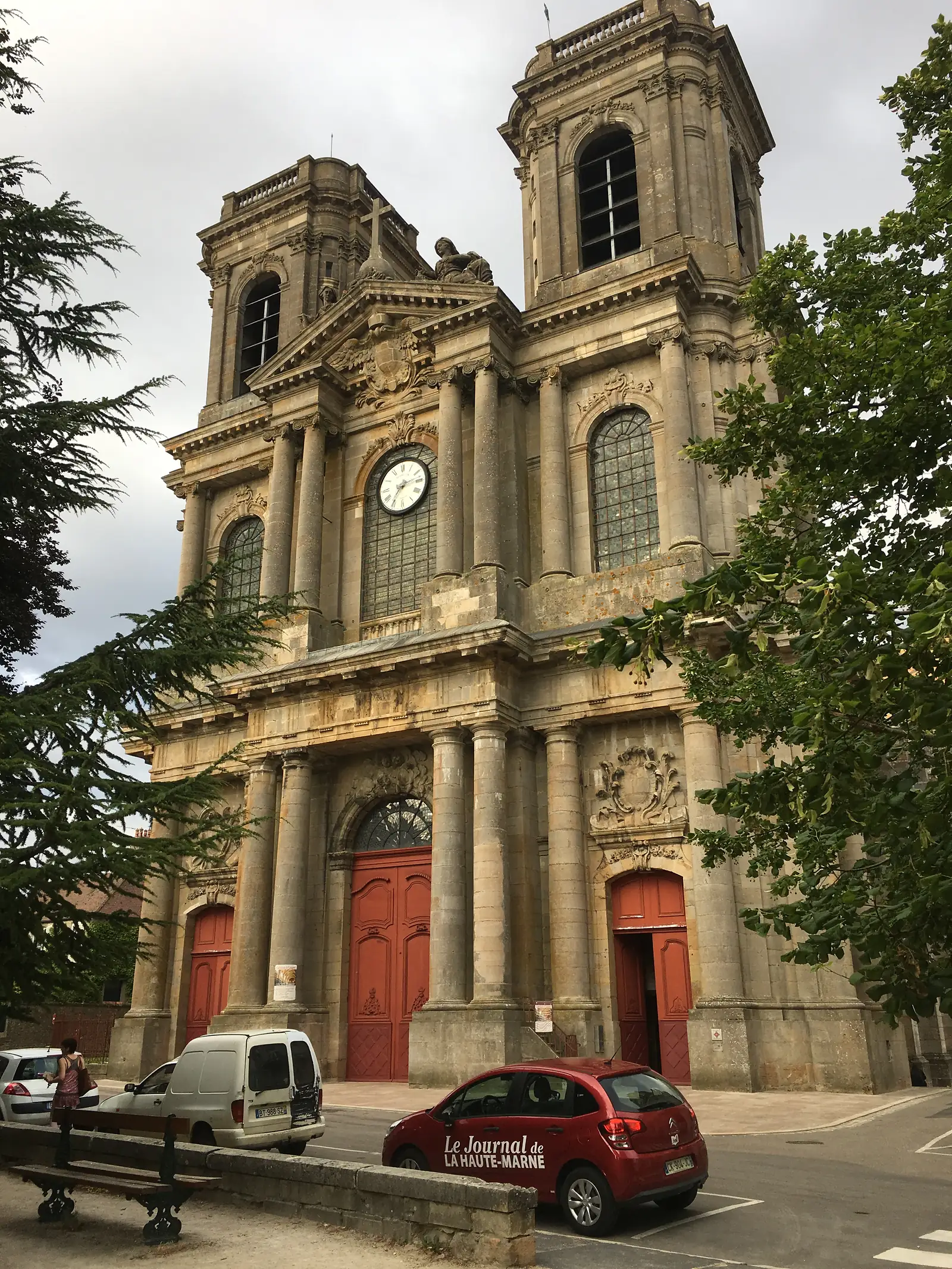The ornate stone facade of a Langres church with a clock tower, red doors and Corinthian pilasters, cars parked outside including one liveried for Le Journal de la Haute-Marne.