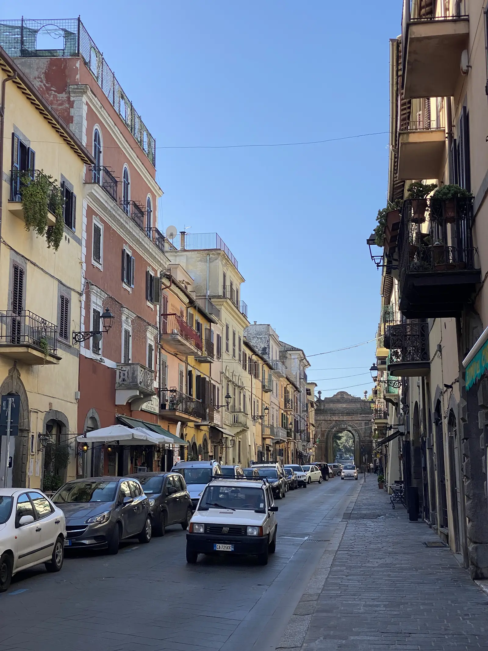 A narrow Italian street lined with tall shuttered townhouses in ochre and terracotta, balconies with potted plants overhead and cars parked along the kerb.