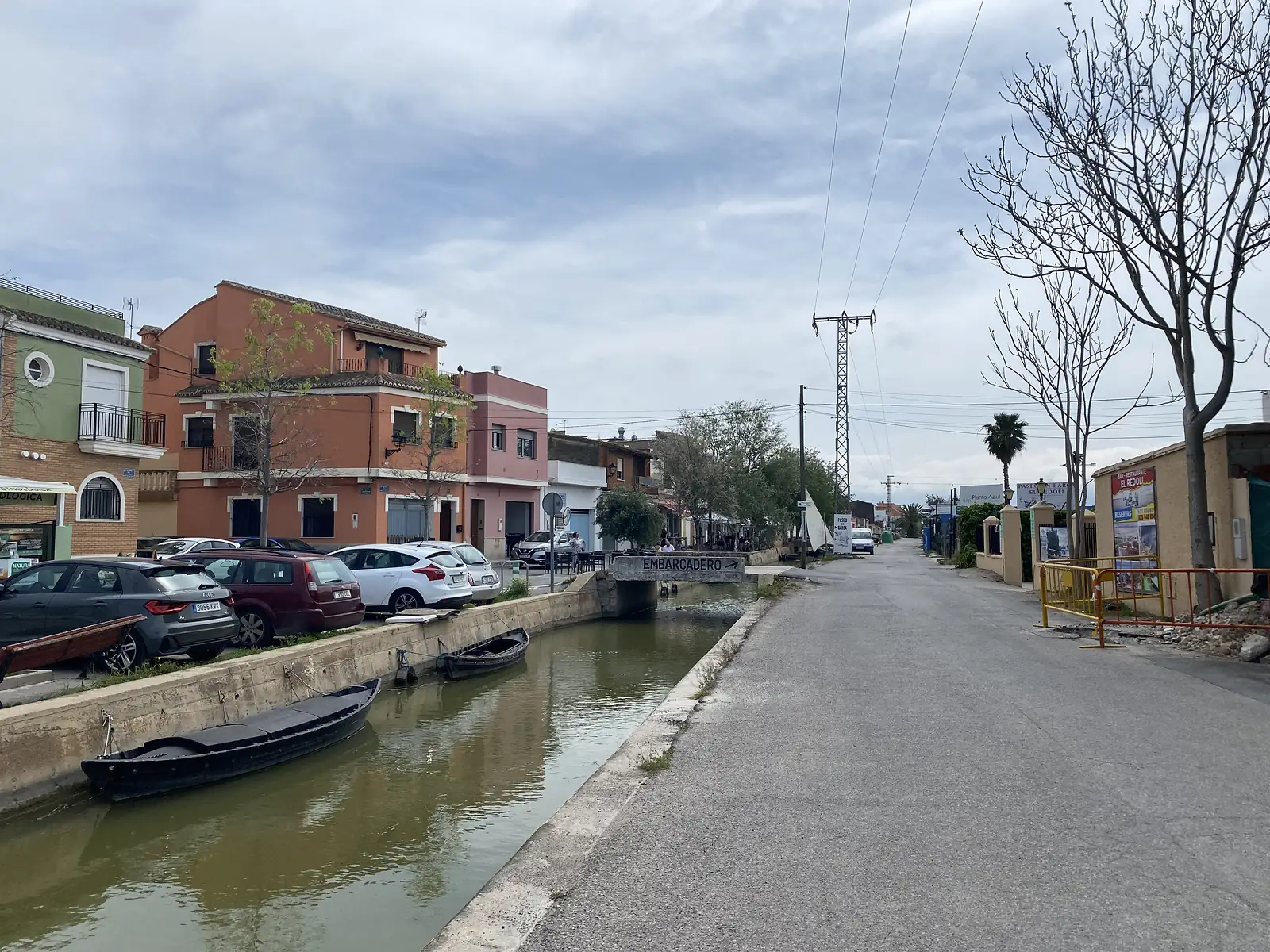 A narrow canal lined with moored black punts running past painted houses, a sign on the bridge reading "EMBARCADERO".