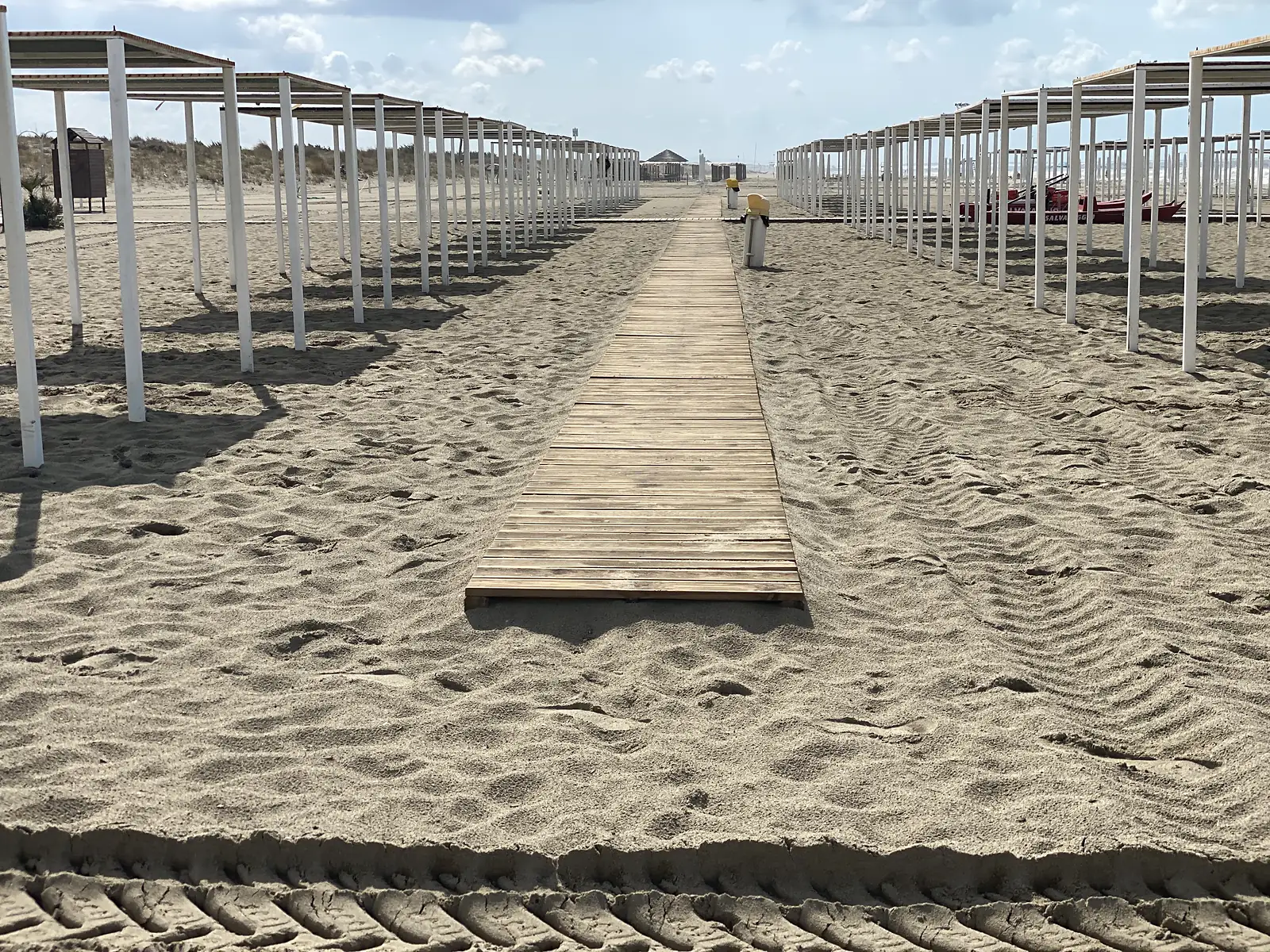 A wooden boardwalk leading through rows of empty beach umbrella frames towards a distant thatched hut on the shore.