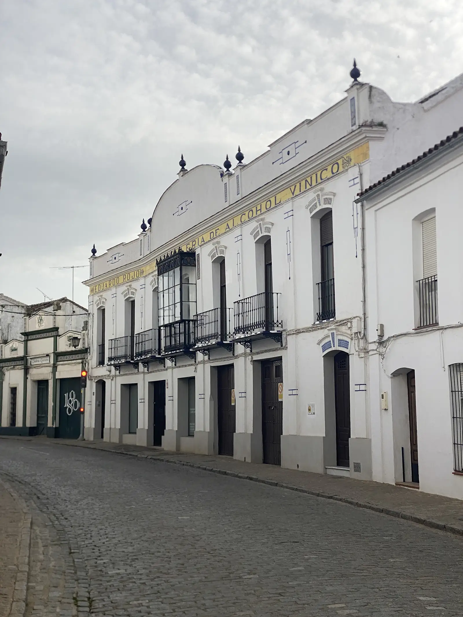 A grand whitewashed building on an empty cobbled street, its front lettered in yellow tiles advertising a wine-alcohol distillery beneath a glazed iron balcony.