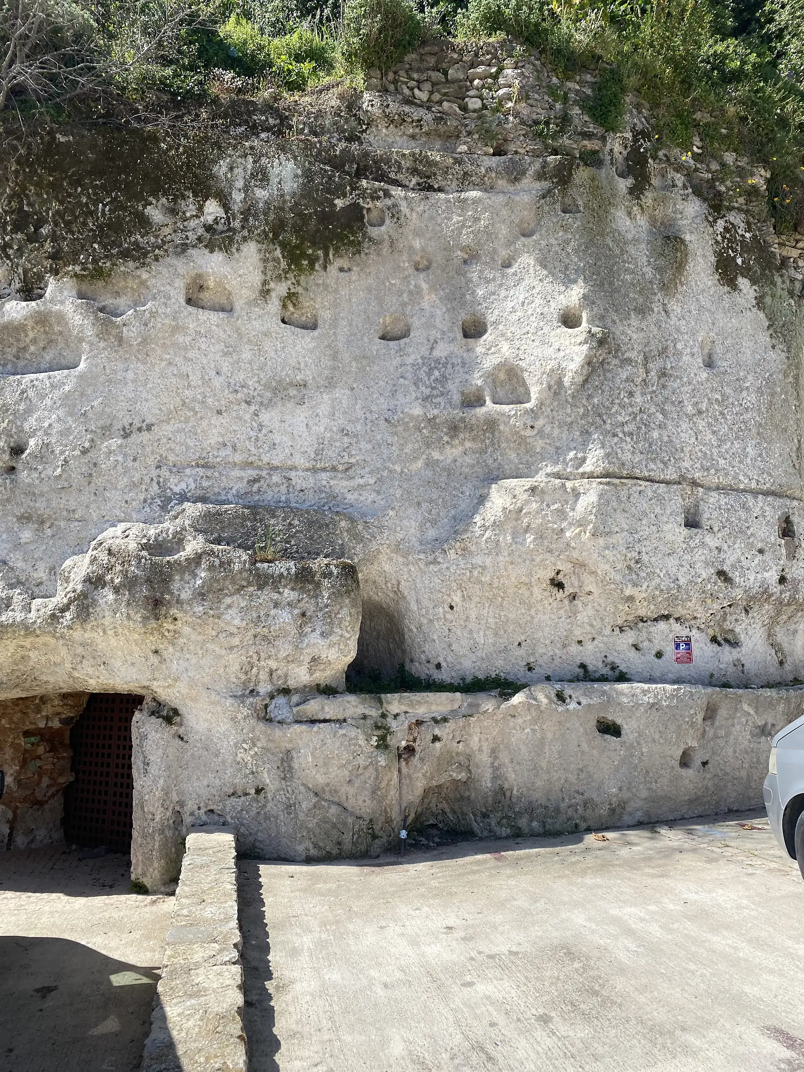 A cliff face pocked with rows of carved hollows and niches above a concrete apron, a lattice-gated cave doorway at its foot.