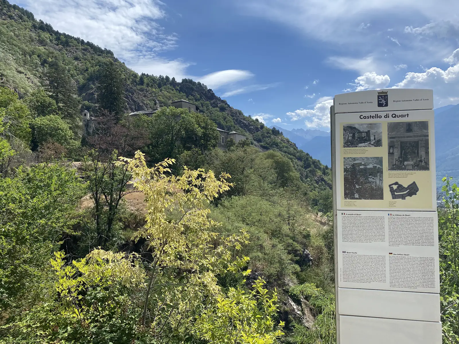 An information panel headed 'Castello di Quart' stands on a wooded hillside path, the castle's towers visible among trees on a rocky spur further along the ridge.