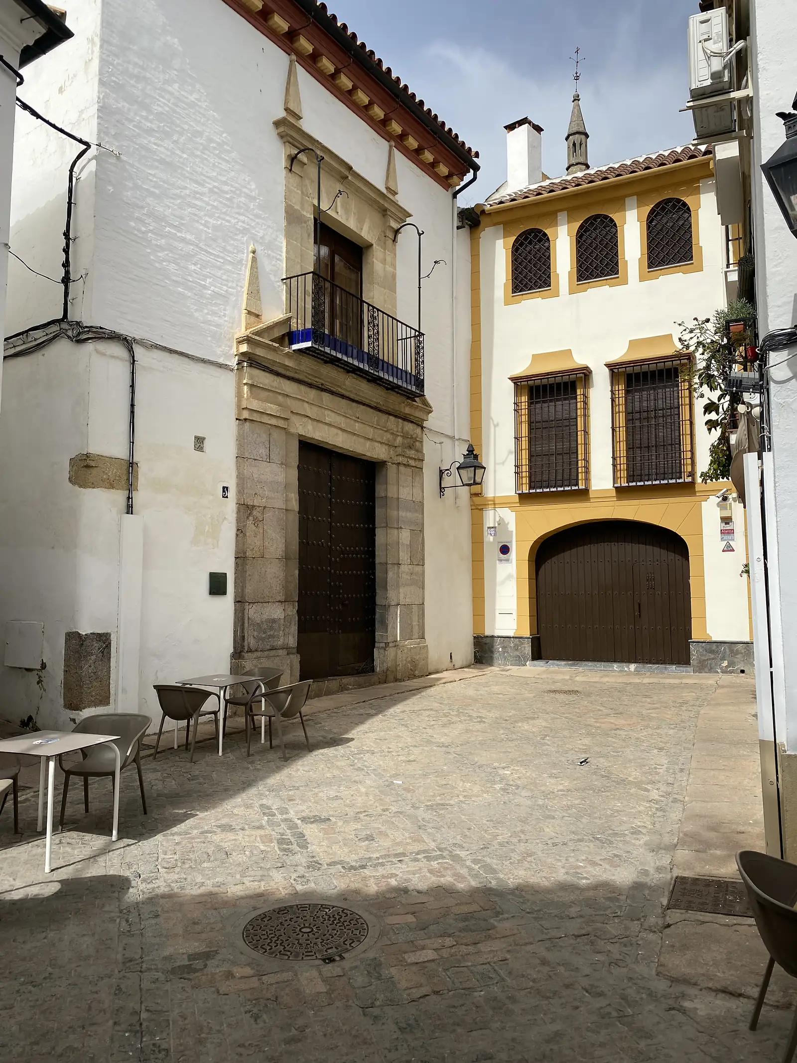Cafe tables set out on a small paved square beside a grand stone doorway, facing a yellow-and-white building with grilled arched windows.