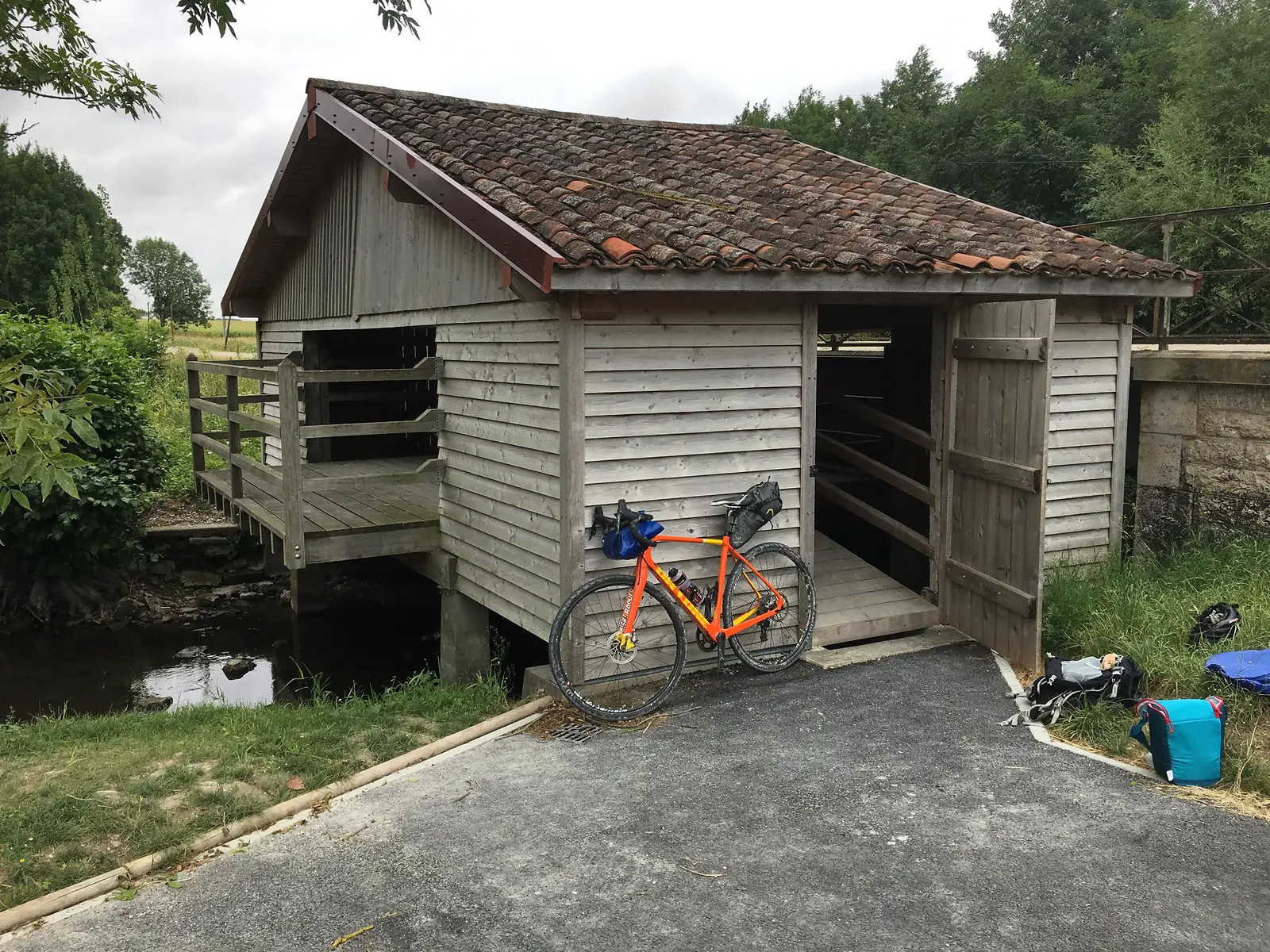 An orange loaded gravel bike leans against a timber-clad pilgrim shelter raised on stilts over a stream, its bags resting nearby on the ground.