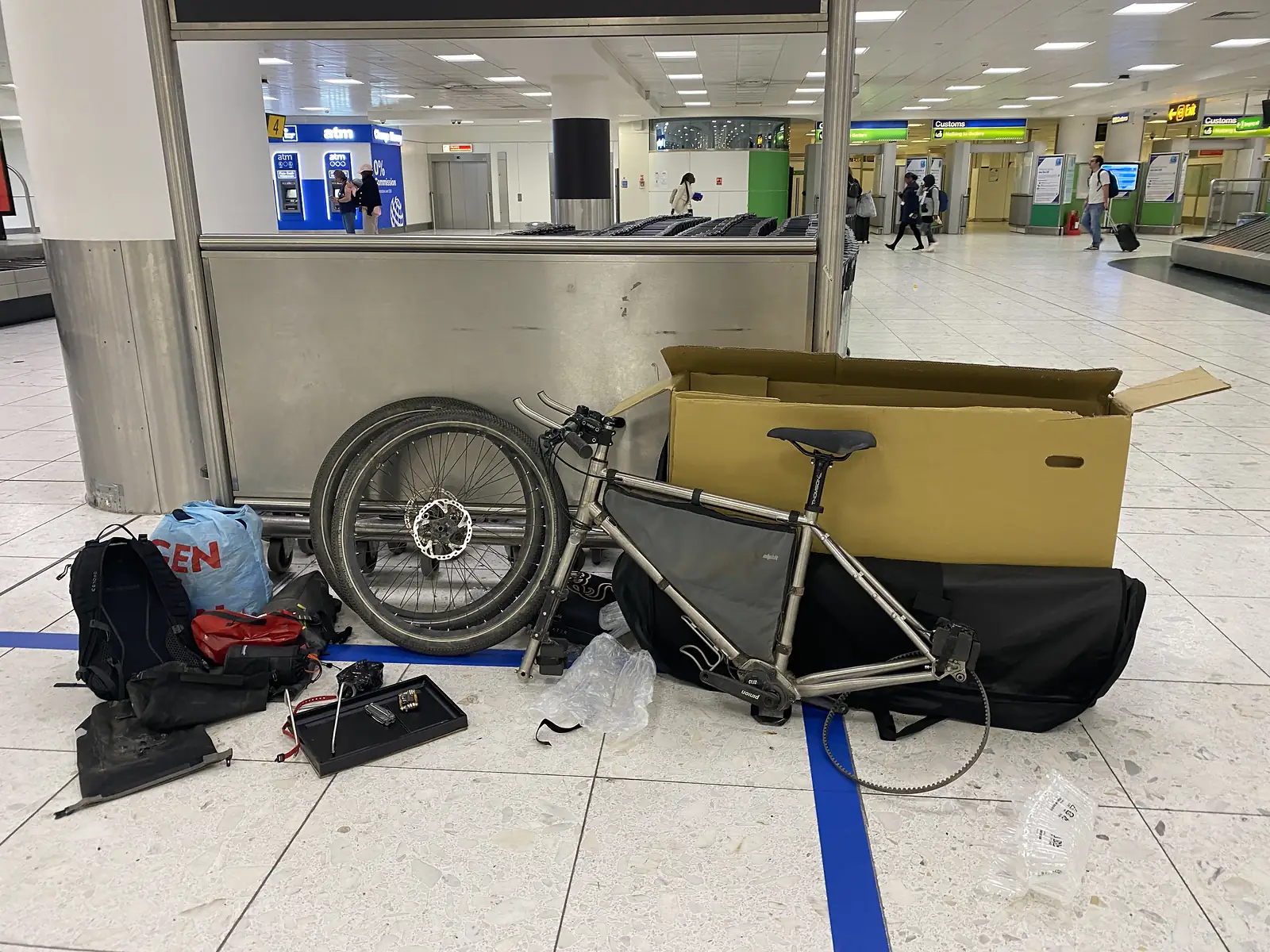 A titanium bike part-dismantled on an airport arrivals floor with its front wheel off, beside a cardboard box, travel bag and rucksack.