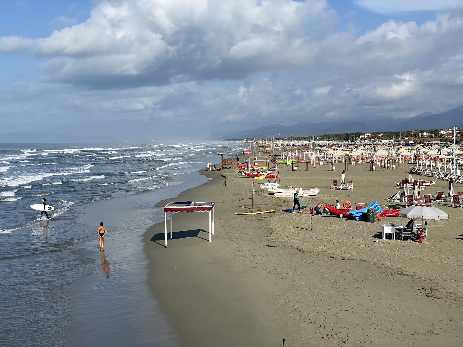A woman in a swimsuit walks along the shoreline past a surfer carrying a board, with beach umbrellas, sunbeds and a lifeguard hut further up the sand.