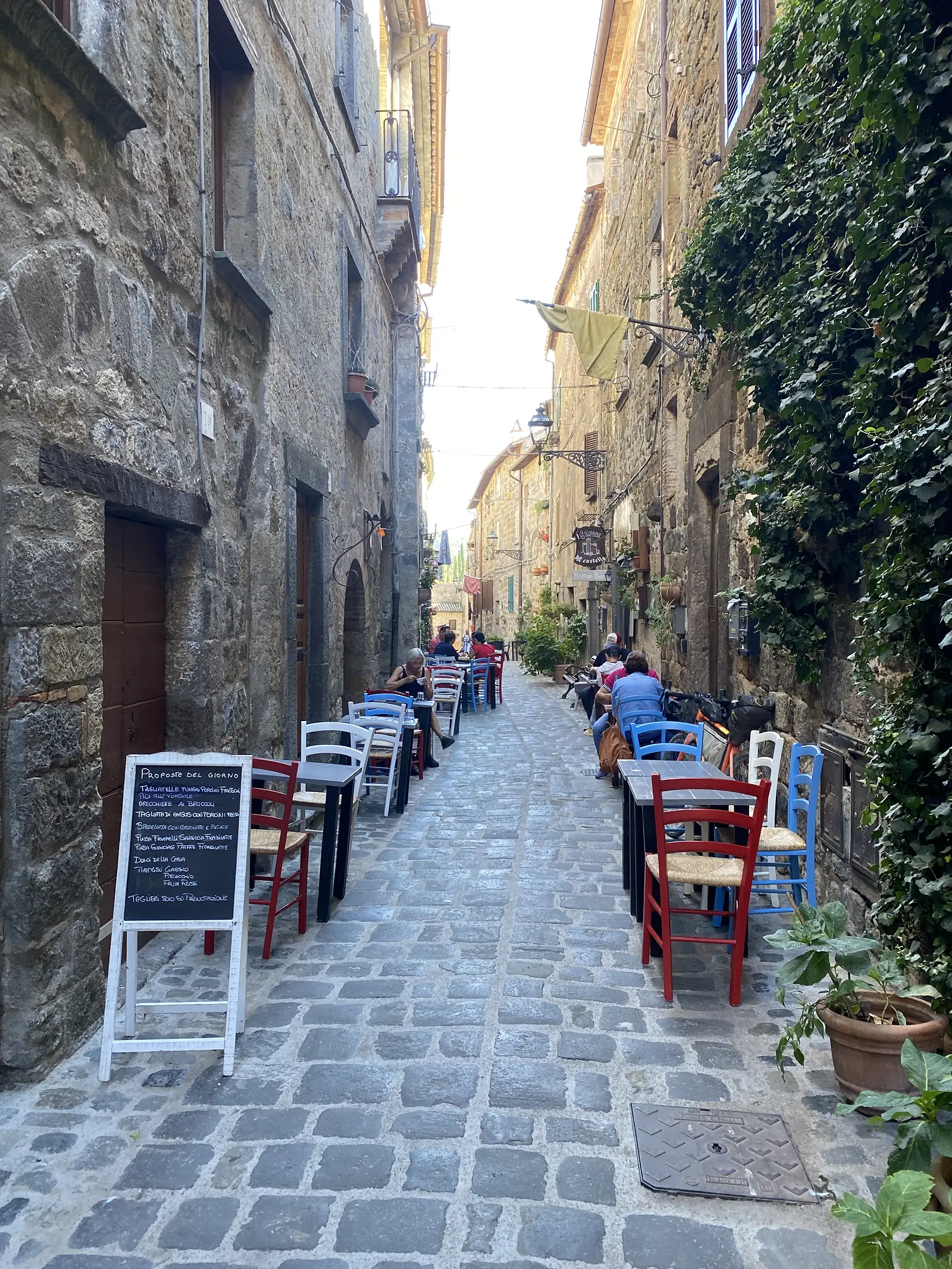 A chalkboard reading "PROPOSTE DEL GIORNO" stands beside colourful cafe chairs on a cobbled street lined with old stone buildings.
