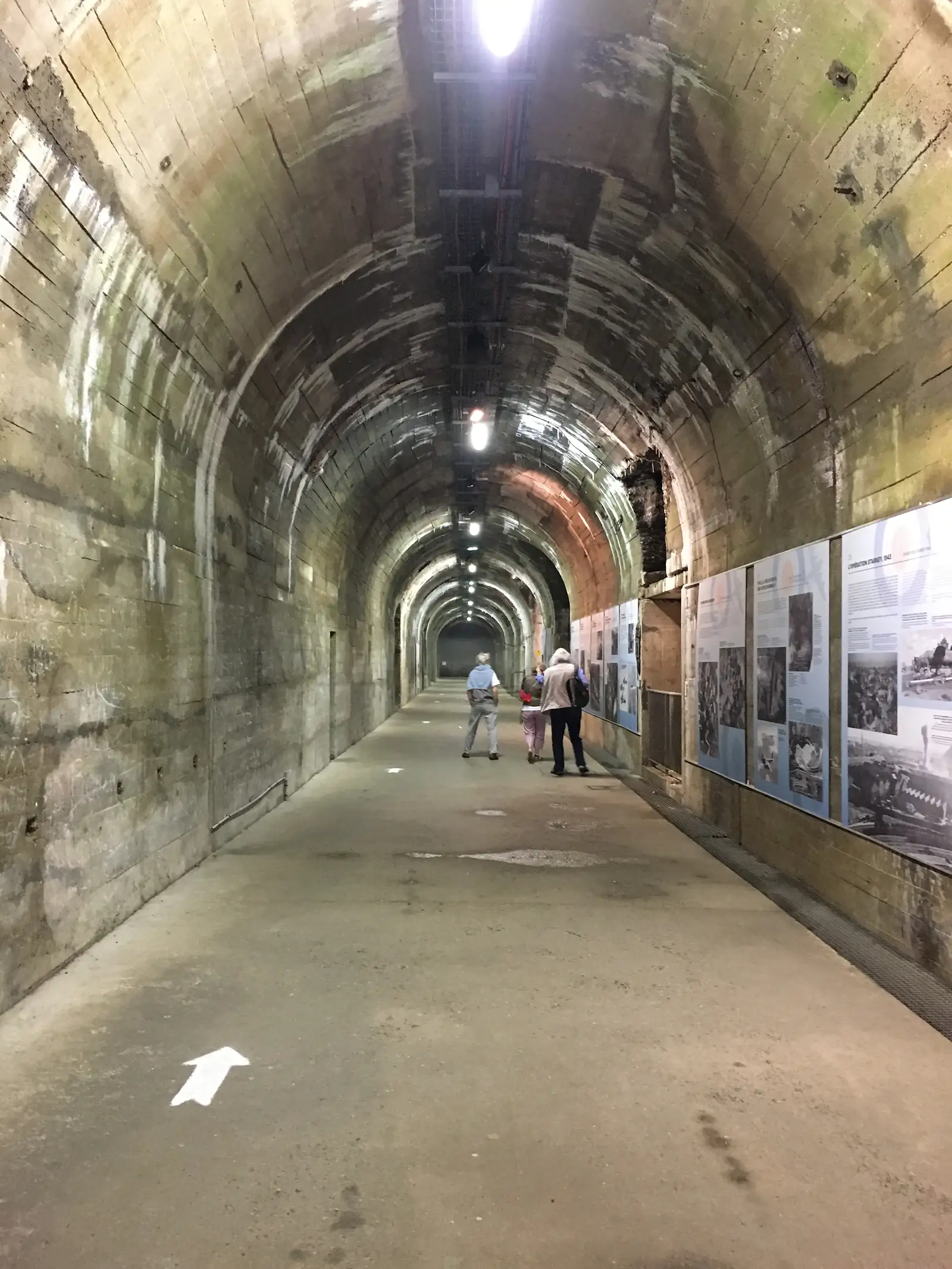 Visitors walk along a long curved tunnel lined with historical information panels overhead, following a white arrow painted on the floor.