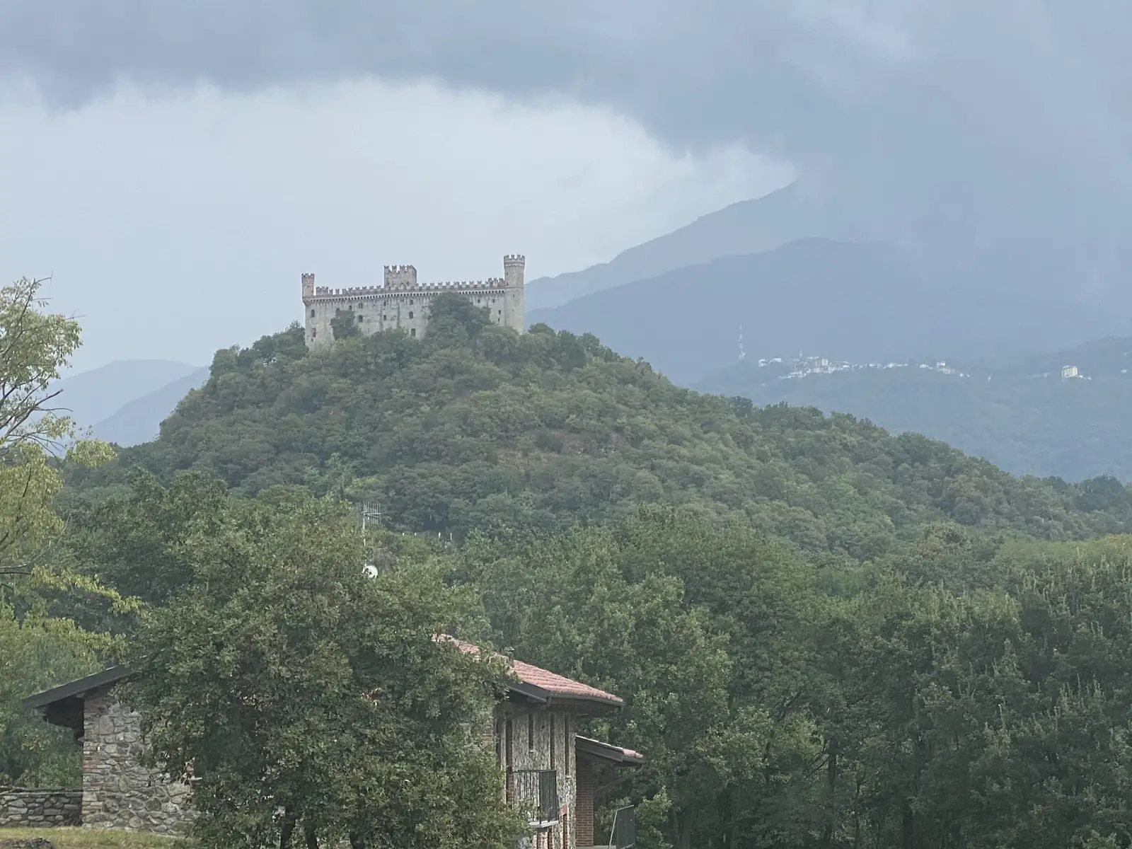 A crenellated stone castle with twin towers sits atop a forested hill under a stormy sky, with a hilltop town visible in the hazy distance towards Ivrea.