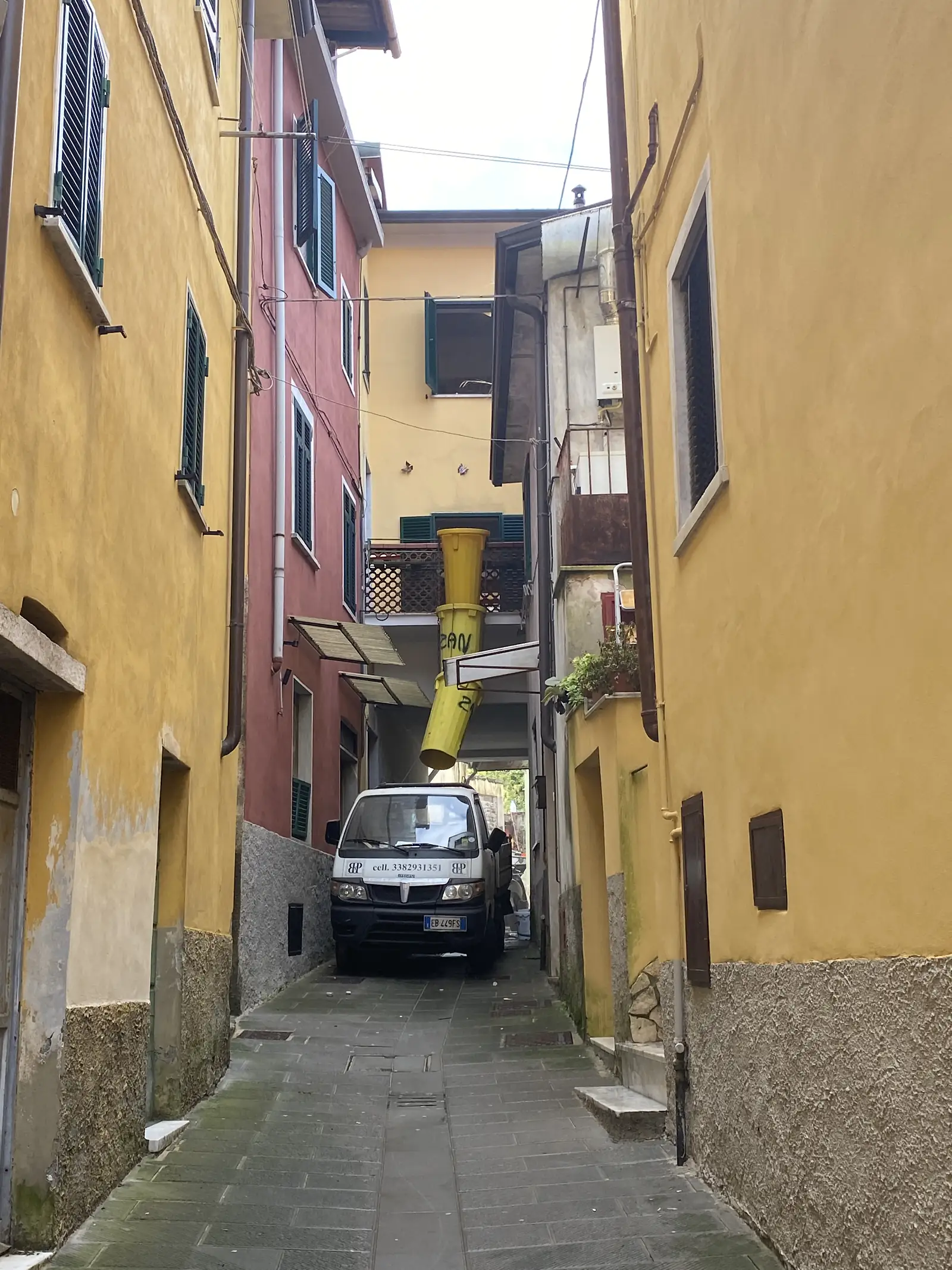 A narrow courtyard between ochre-rendered buildings, with a small van parked beside a yellow builder's rubble chute running down from an upper-storey window.
