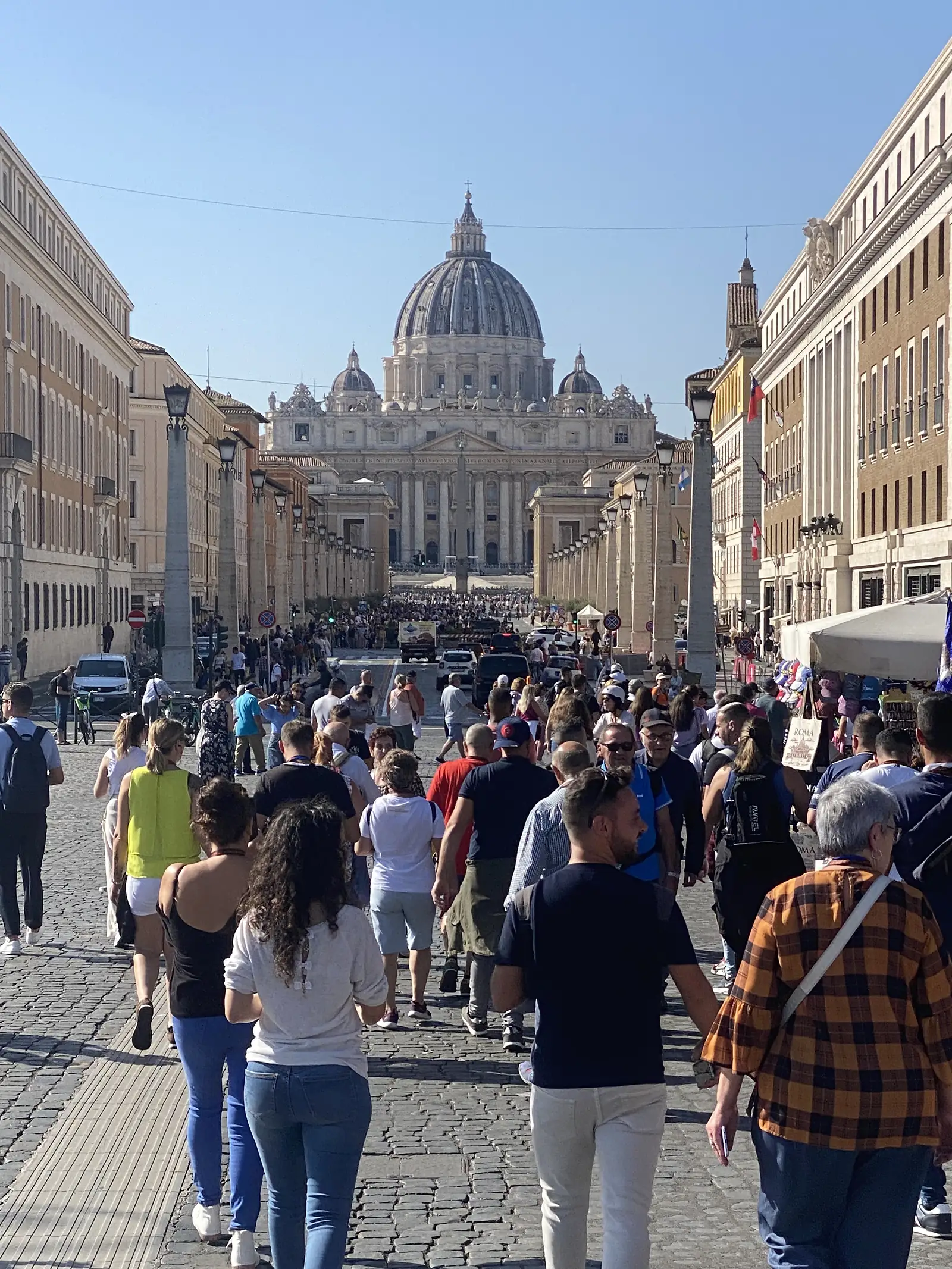 Crowds walk down a wide boulevard towards the dome of St Peter's Basilica rising above Rome's rooftops in the distance.