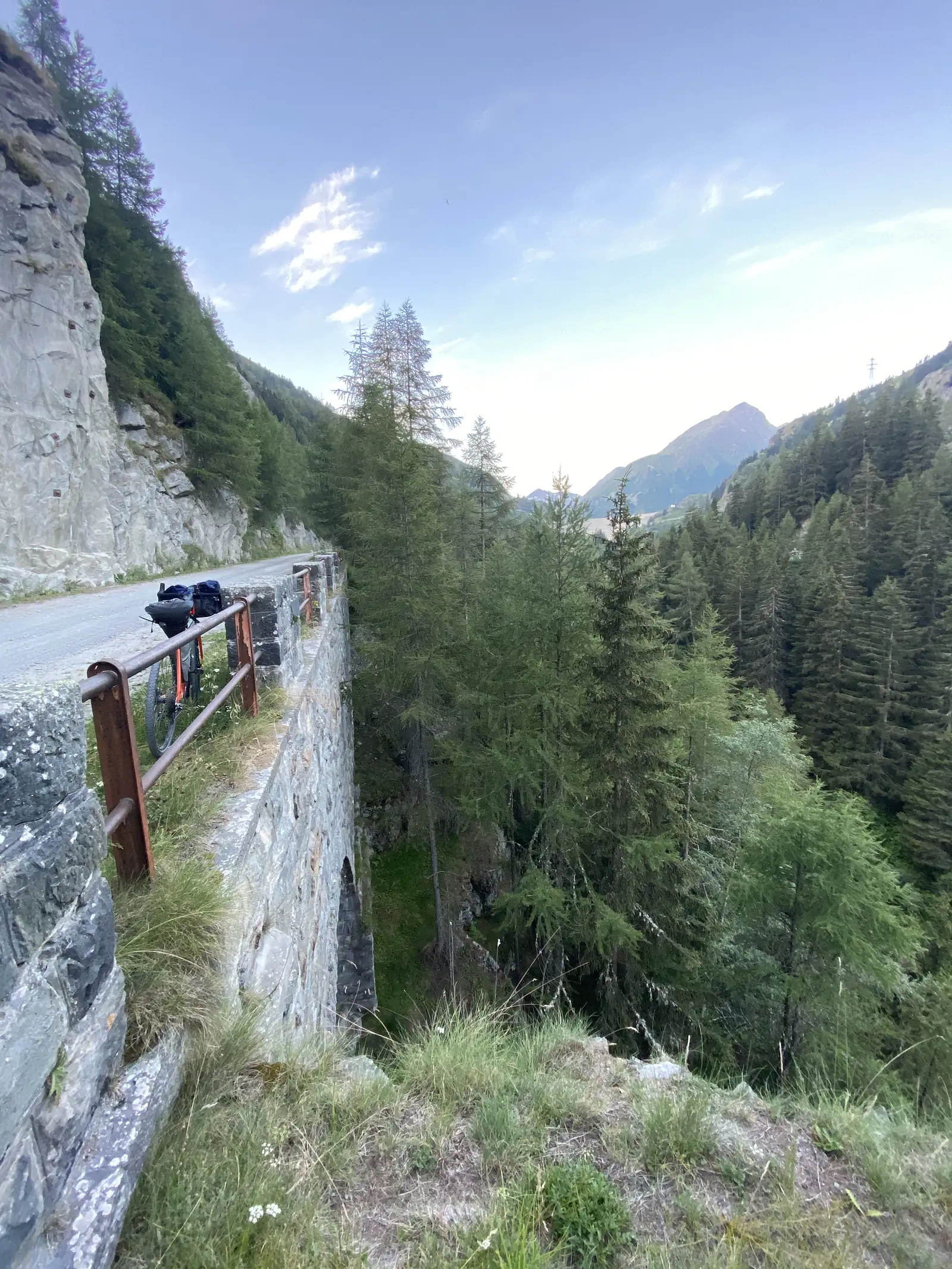 A loaded touring bike leans against the stone parapet of an old railway viaduct spanning a wooded ravine, a mountain peak visible down the valley.