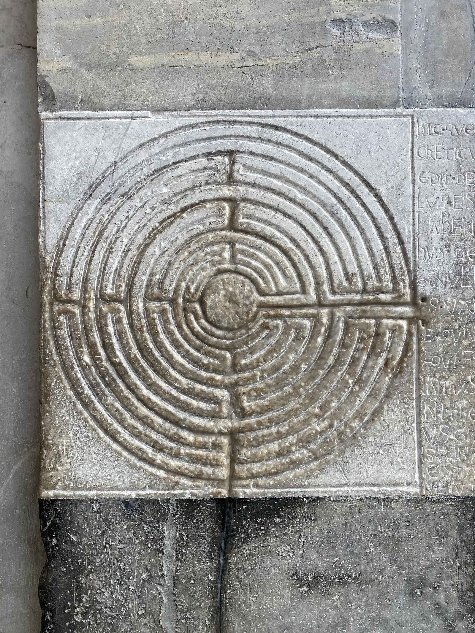 A stone relief carving of a circular seven-ring labyrinth set into a cathedral wall, surrounded by faint Latin inscriptions in worn capital lettering.
