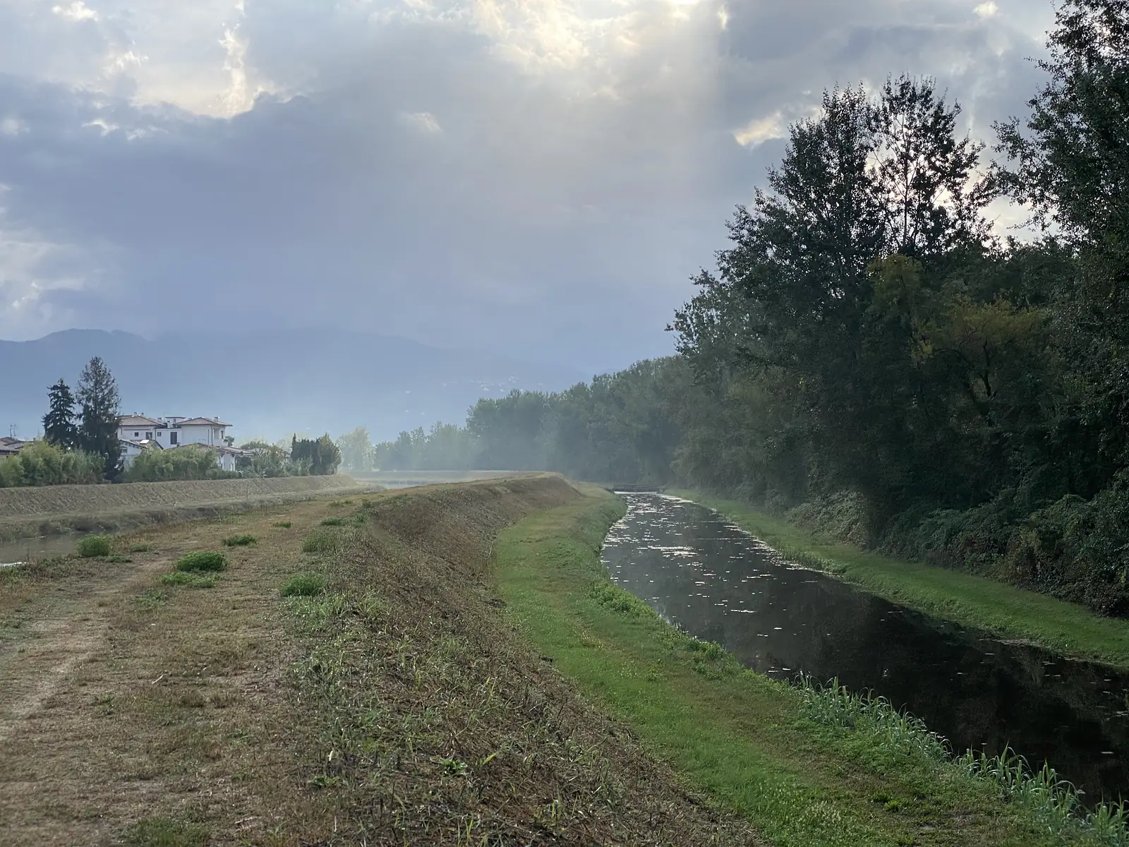 A grassy flood-defence bank runs alongside a still, tree-lined drainage canal under a misty morning sky, with hazy hills visible in the distance.