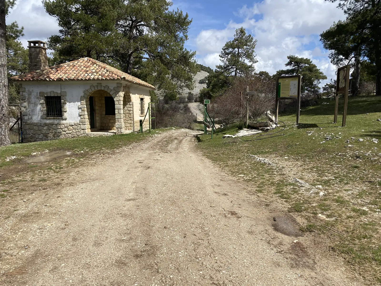 A small stone and white forest lodge with an arched porch beside a track junction, with wooden information boards and a signpost.