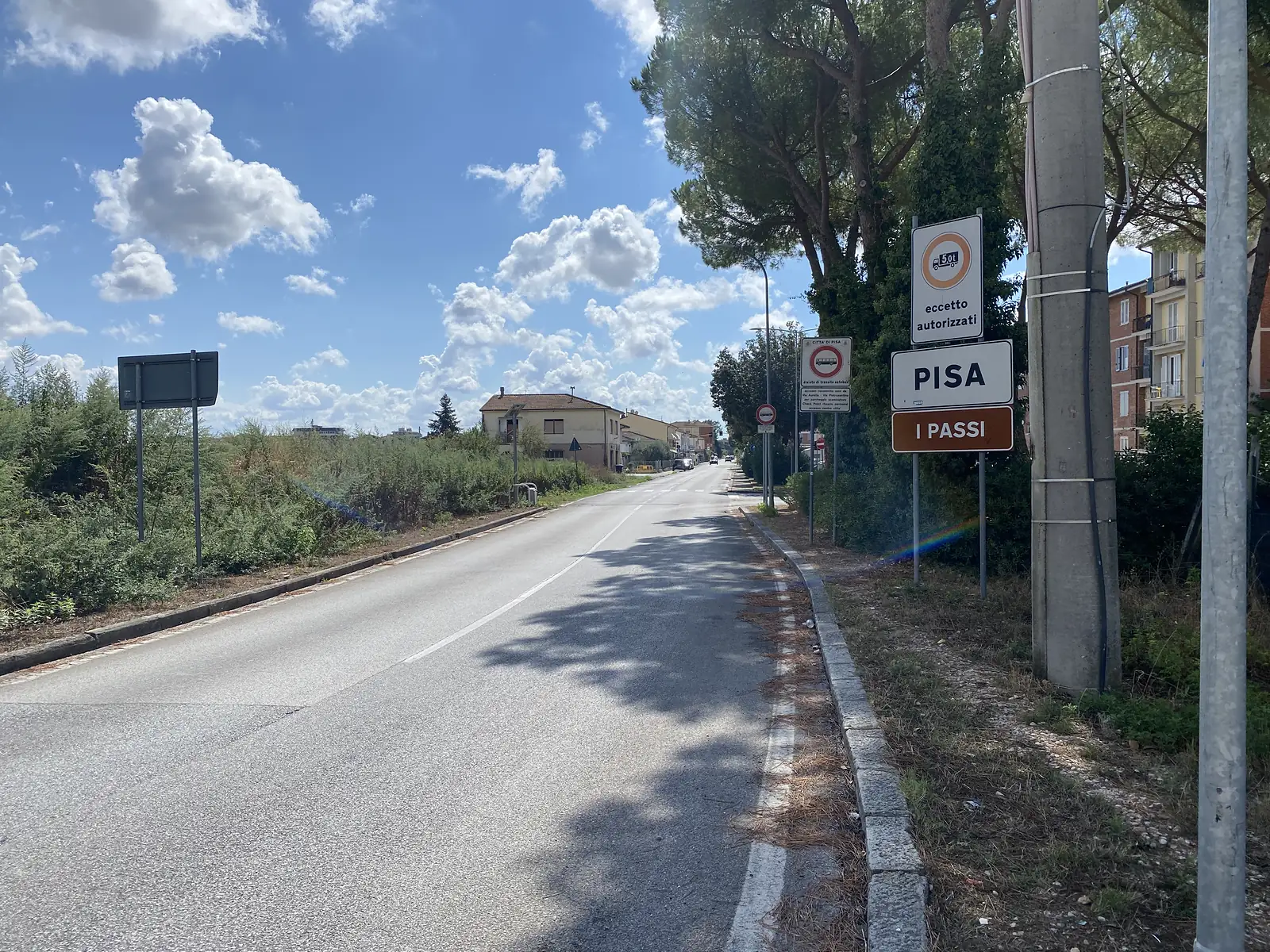 A road sign reading Pisa and I Passi marking the town boundary, with a tree-lined street continuing ahead.