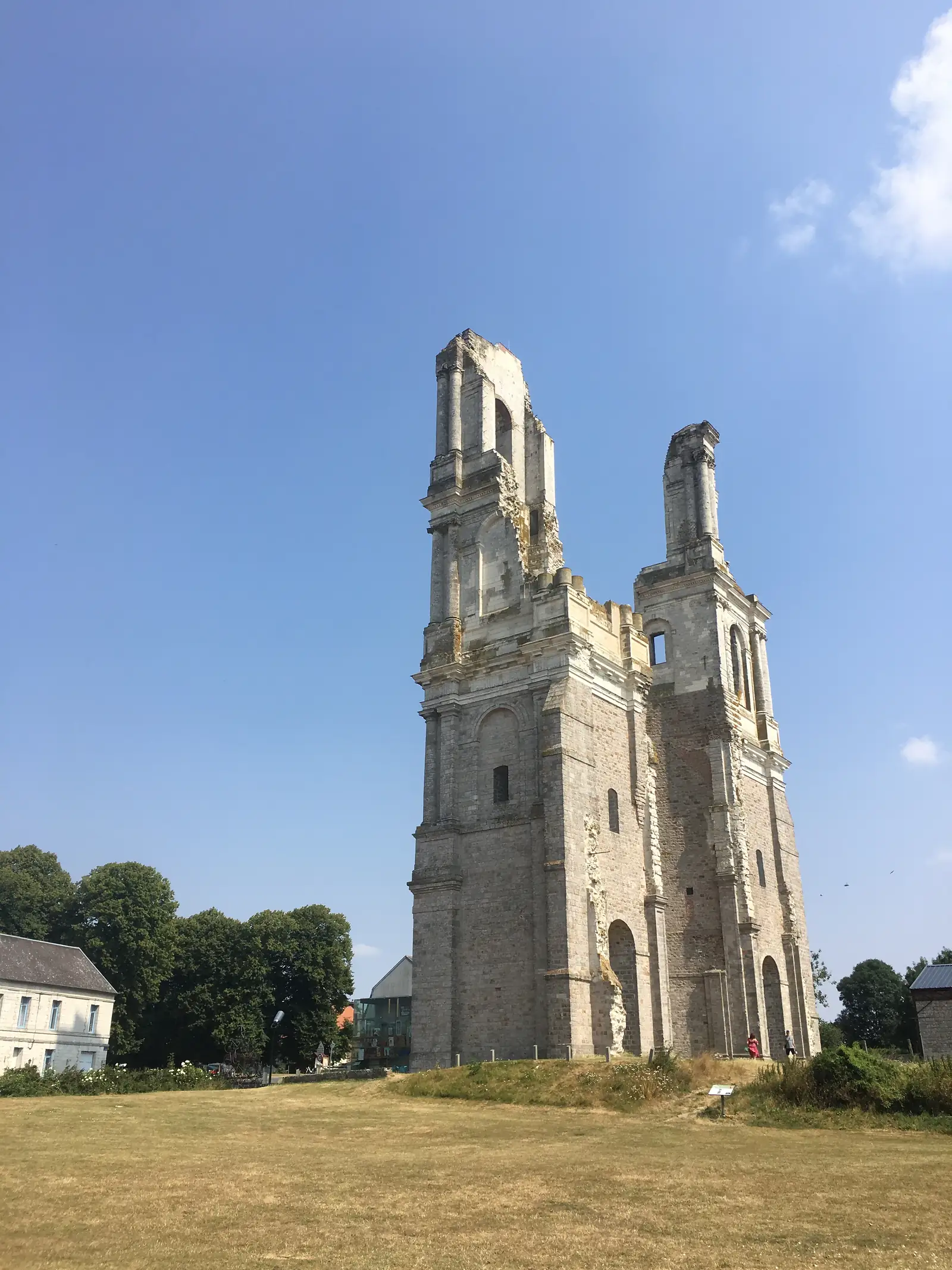 A ruined brick church with a soaring spire and empty tracery windows, its roof gone, standing beside mature trees under a clear sky.