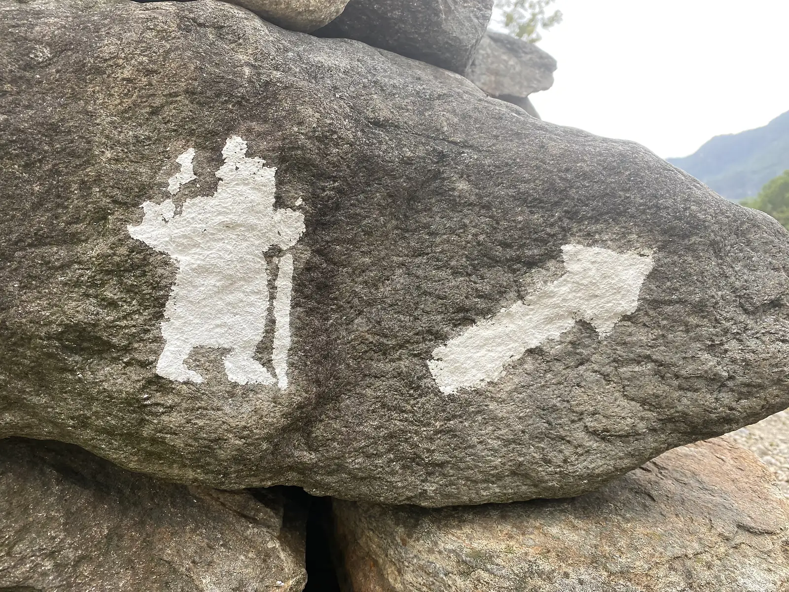 A white pilgrim-and-scallop waymark and directional arrow are painted onto a large boulder, marking a Via Francigena route.