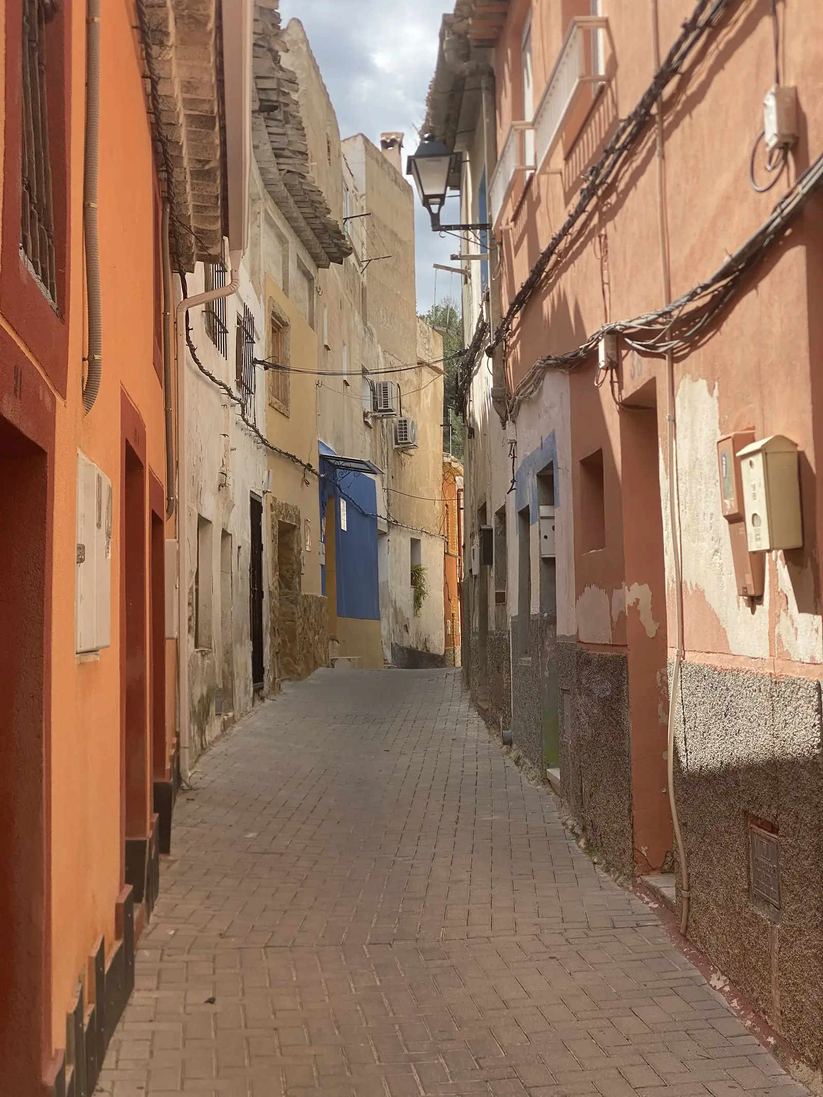 A narrow brick-paved alley climbing between tall houses rendered orange, ochre and blue, with tangled cables strung overhead and an iron lamp.