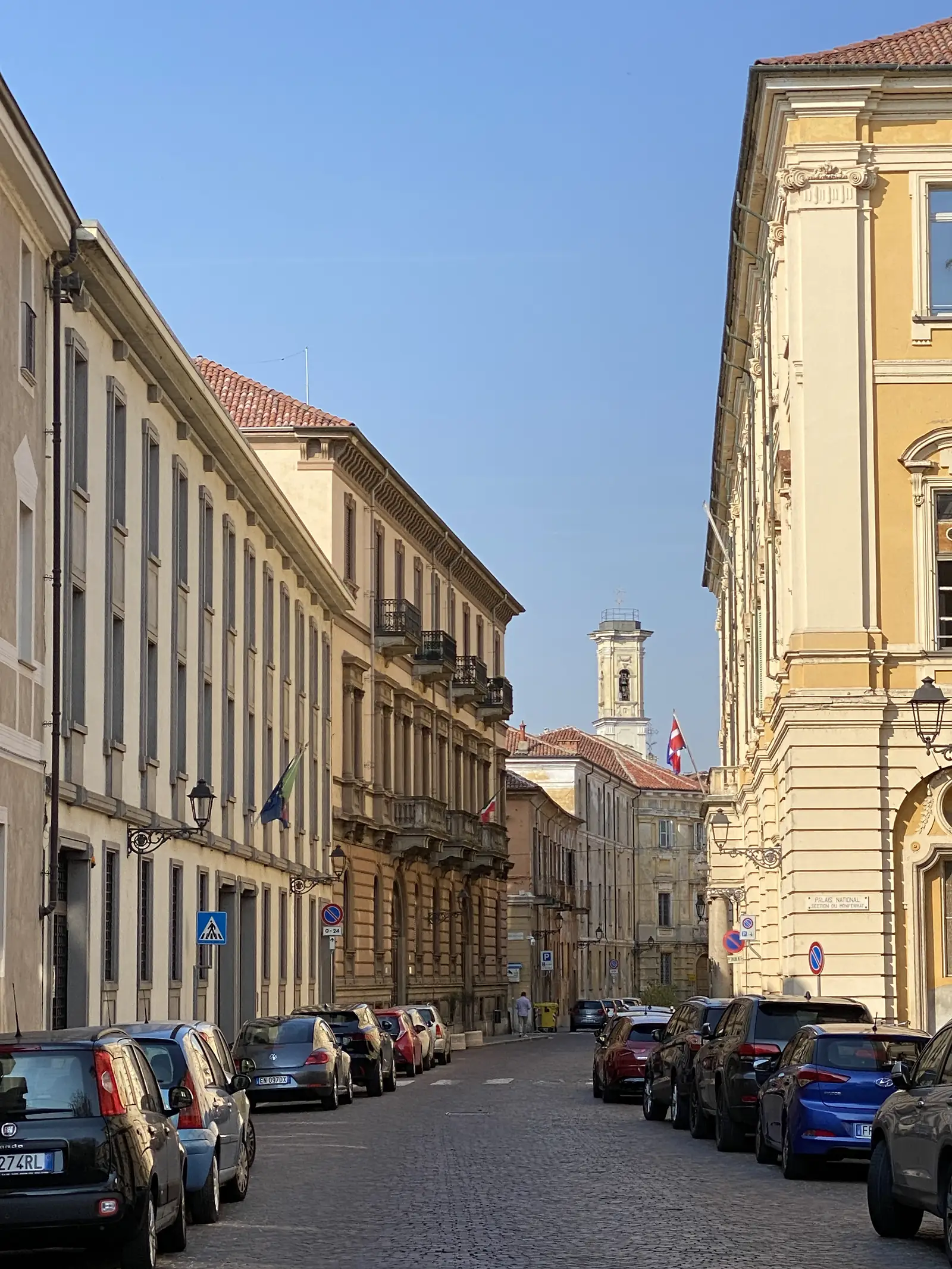 A colonnaded arcade along an Italian piazza, flags hanging from the upper storeys and a stone clock tower rising above the rooftops beyond.