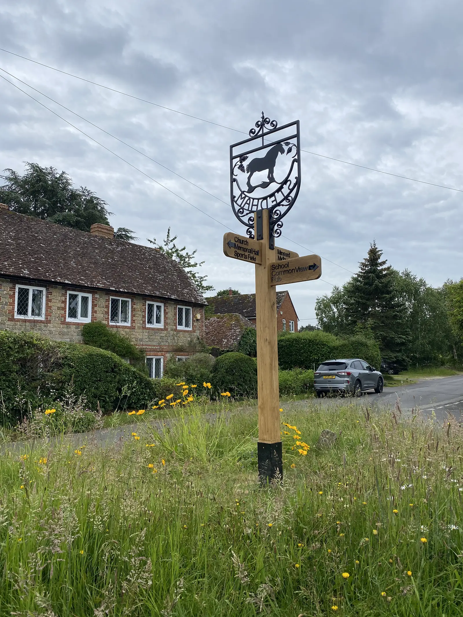 A wrought-iron village sign showing a shire horse, on a post with fingerposts to the church, memorial hall, school and pub, in a verge of tall grass.