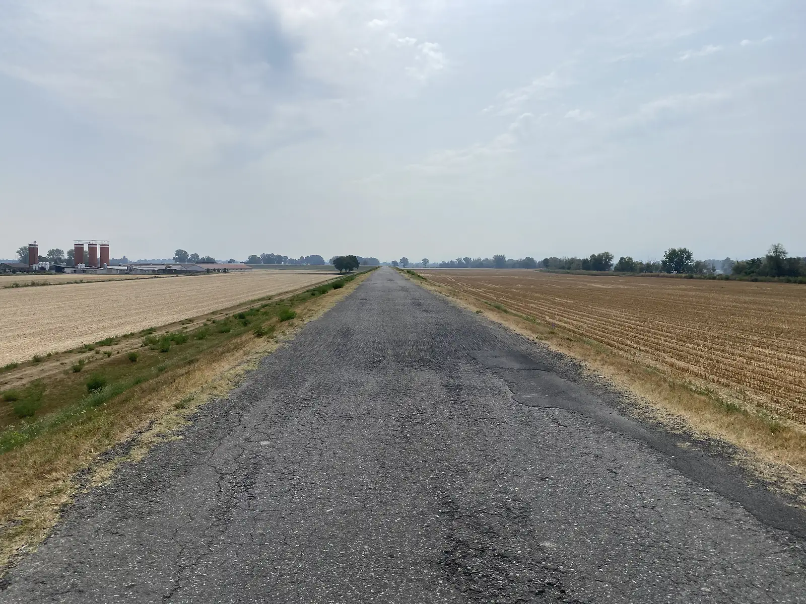 A long, cracked tarmac road runs dead straight between stubbled fields towards farm buildings and grain silos on the horizon.