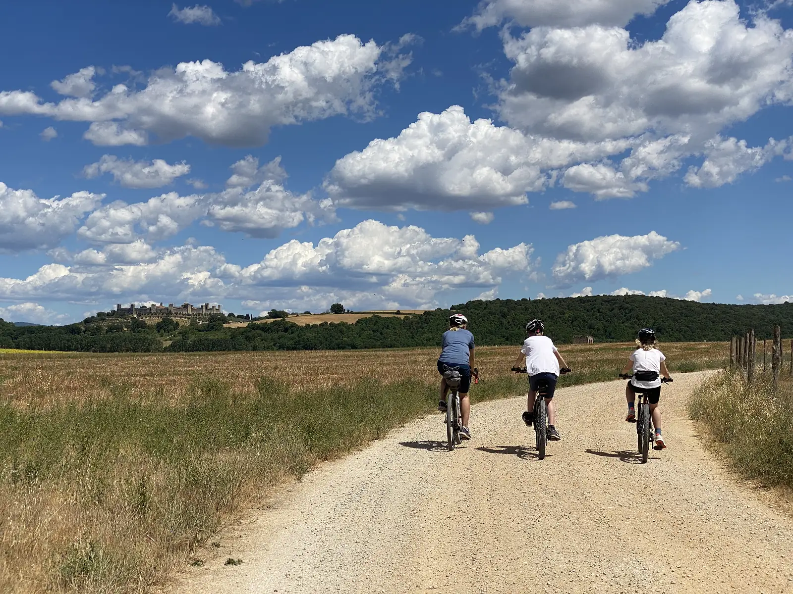 Three cyclists ride away along a white gravel road between dry fields under a big sky of cumulus clouds, a walled hill town visible far ahead.
