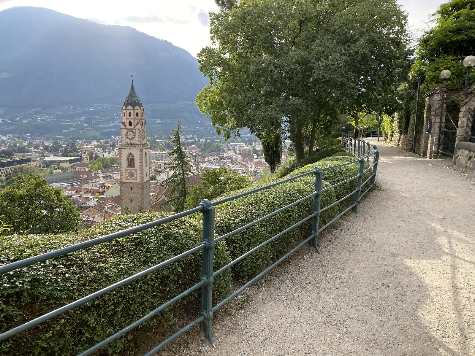 A gravel promenade runs behind a green railing and clipped hedge high above a town, with a tall clock tower below and a mountain flank behind.