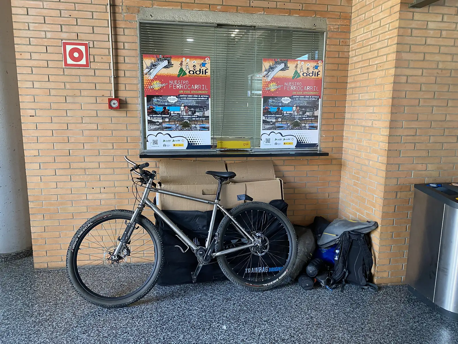 A titanium mountain bike rebuilt and leaning against a brick station wall beside its empty padded travel bag, luggage and a rucksack.